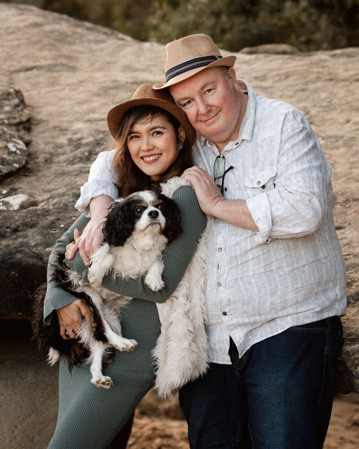 A couple poses together on rocky terrain with their small Cavalier King Charles Spaniel dog  in sweet portraits in Rouse Hill Photoshoot