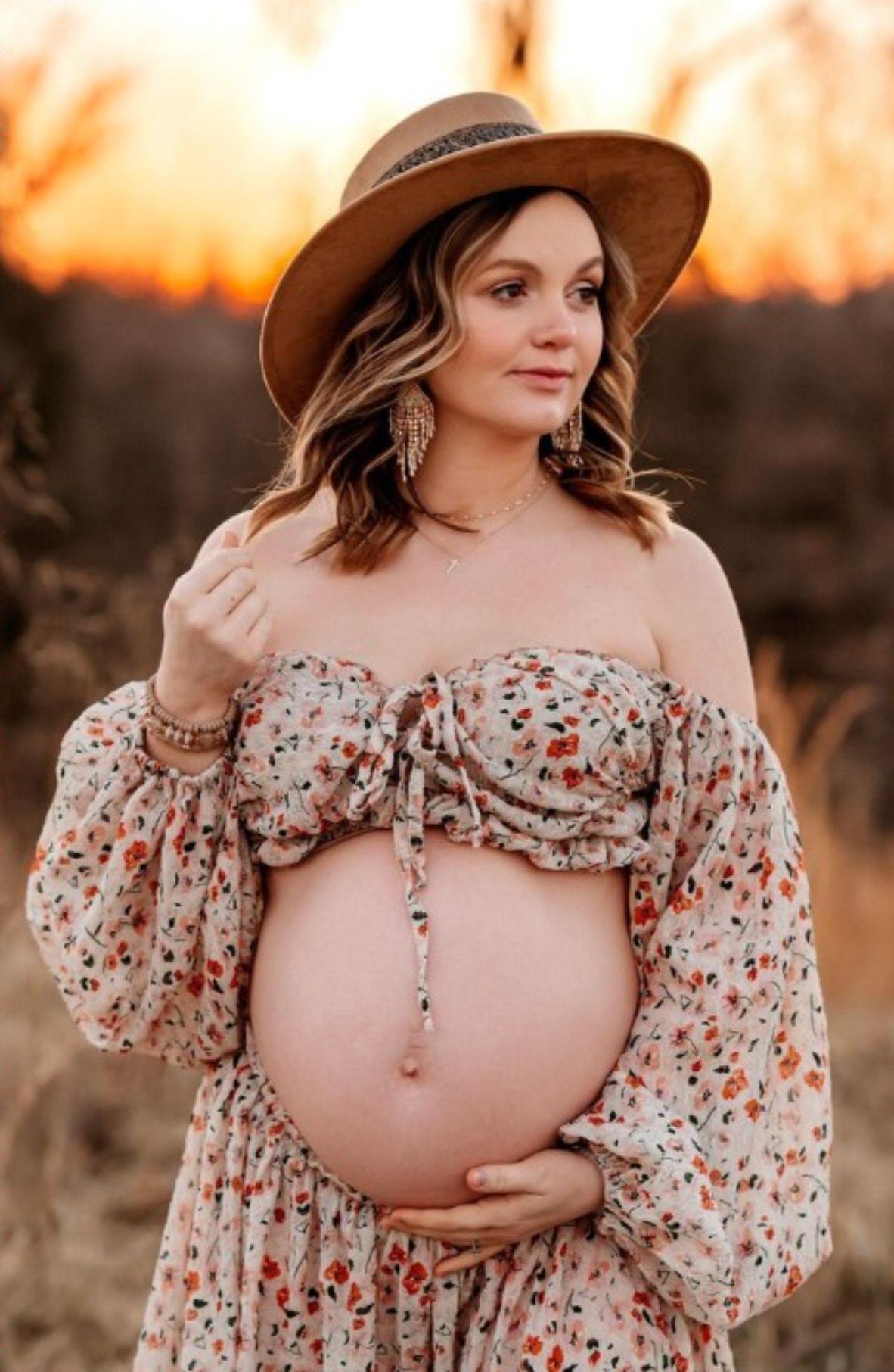 A pregnant woman in a floral dress and wide brim hat poses in a desert landscape at sunset.