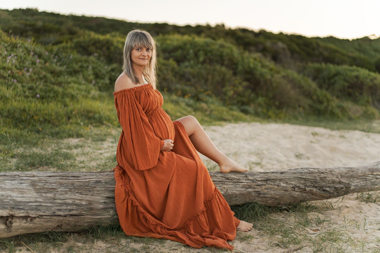 A pregnant woman in a flowing rust-colored dress sits gracefully on driftwood at a scenic beach during golden hour maternity photoshoot