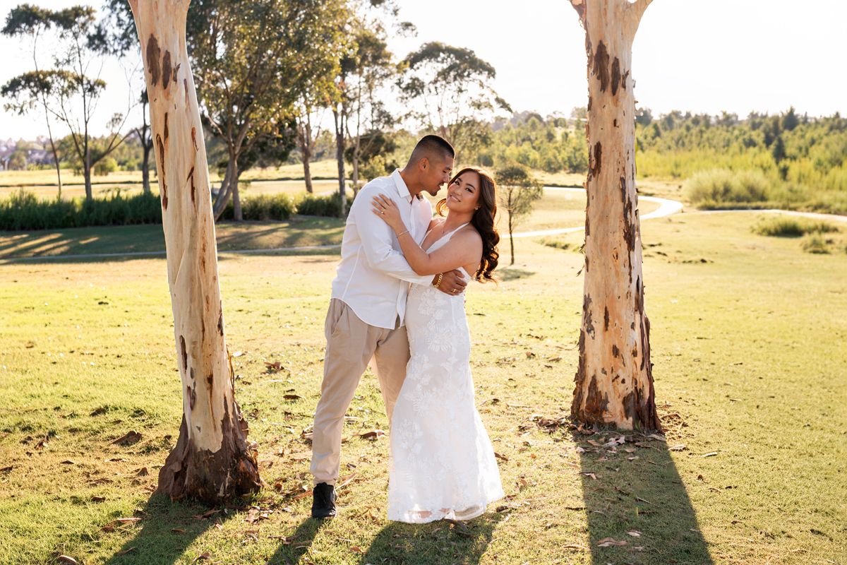 A couple in white attire share a romantic moment between tall eucalyptus trees in a sunlit outdoor setting during an elopement photoshoot