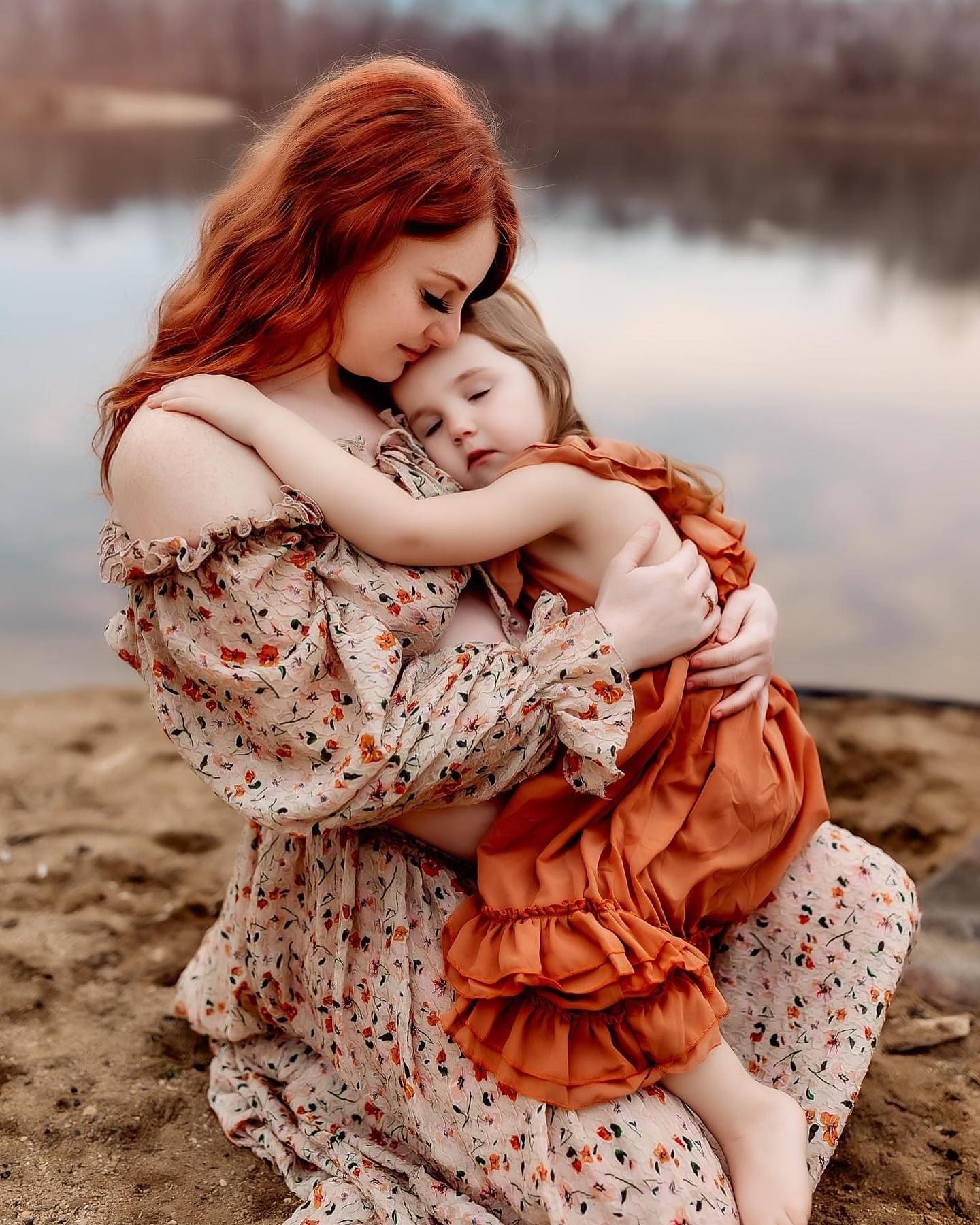 A tender moment captured at the beach with floral and rust-colored dresses against a serene waterfront backdrop.
