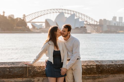 A couple shares a romantic moment by Sydney Harbour with the Opera House and Harbour Bridge in the background. Engagement photoshoot Sydney