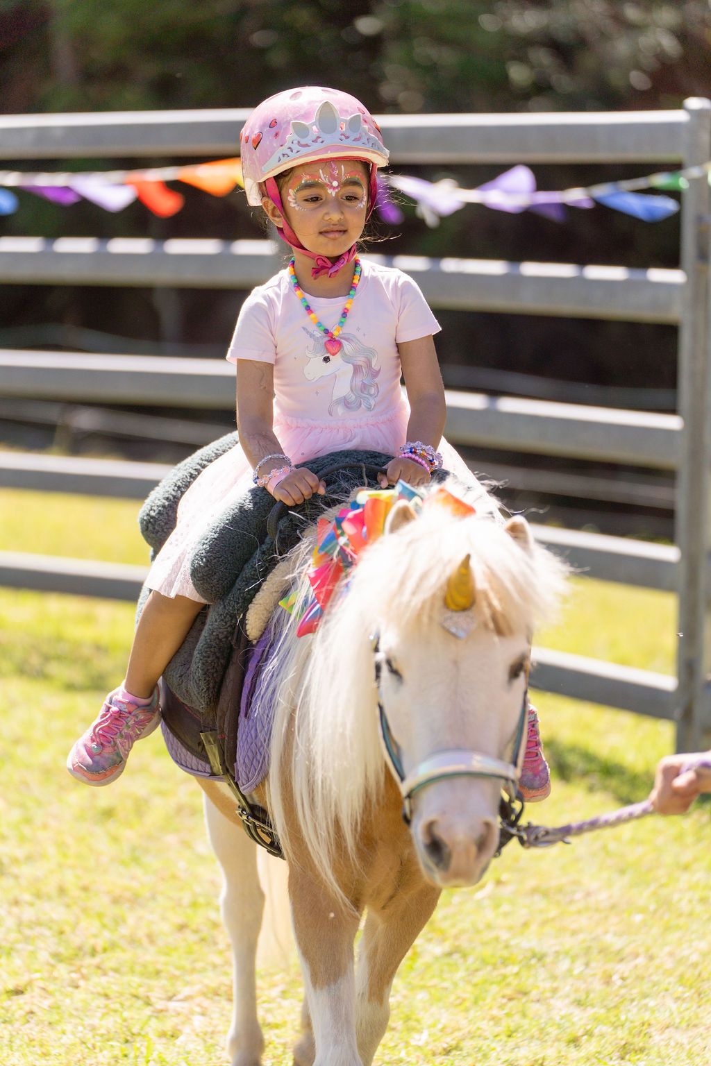 A small child rides a white pony wearing pink attire during a sunny outdoor event and a photographer captures it