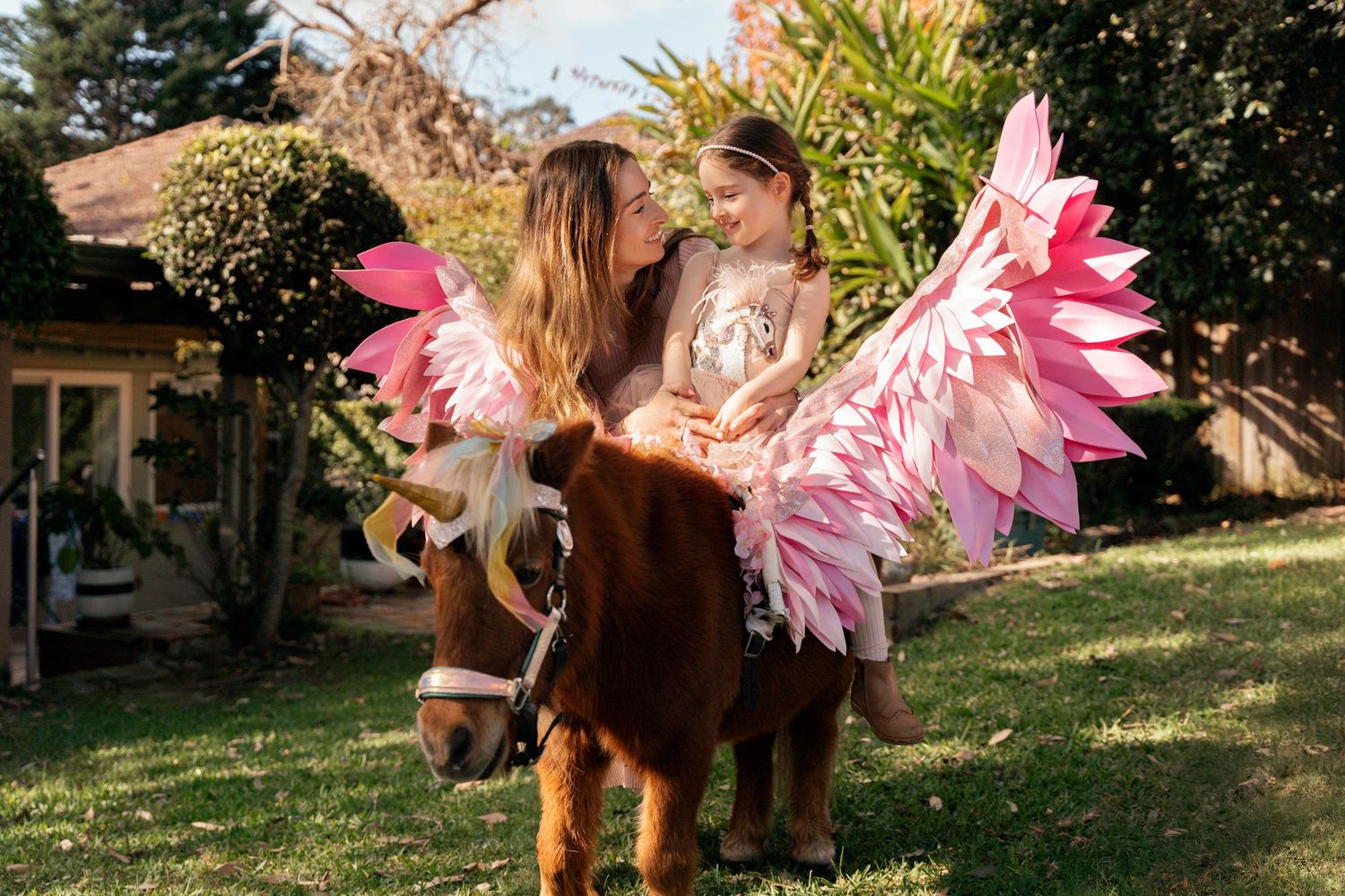 A brown pony toy with pink fairy wings stands in a sunny garden setting while mom looks at her daughter during a photoshoot with a pony