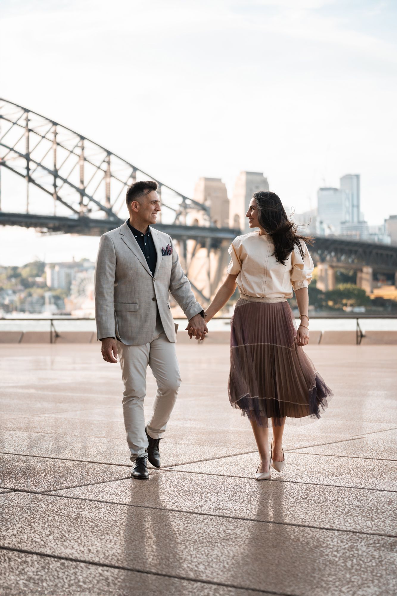 Couple holding hands and walking along Sydney Harbour with the iconic bridge in the background during sunset.