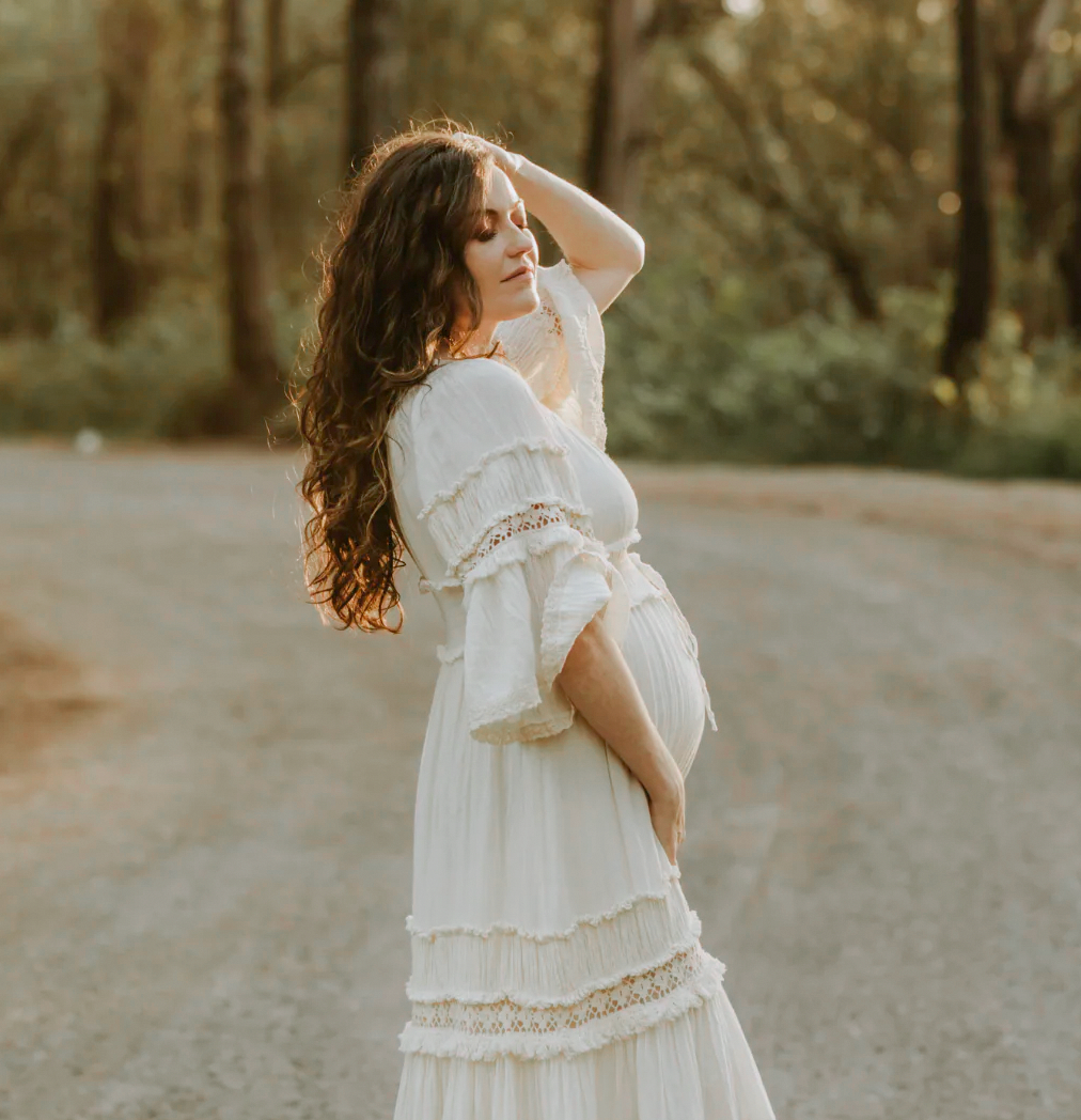 Woman in flowing white lace dress poses on dirt road during golden hour in forest setting.
