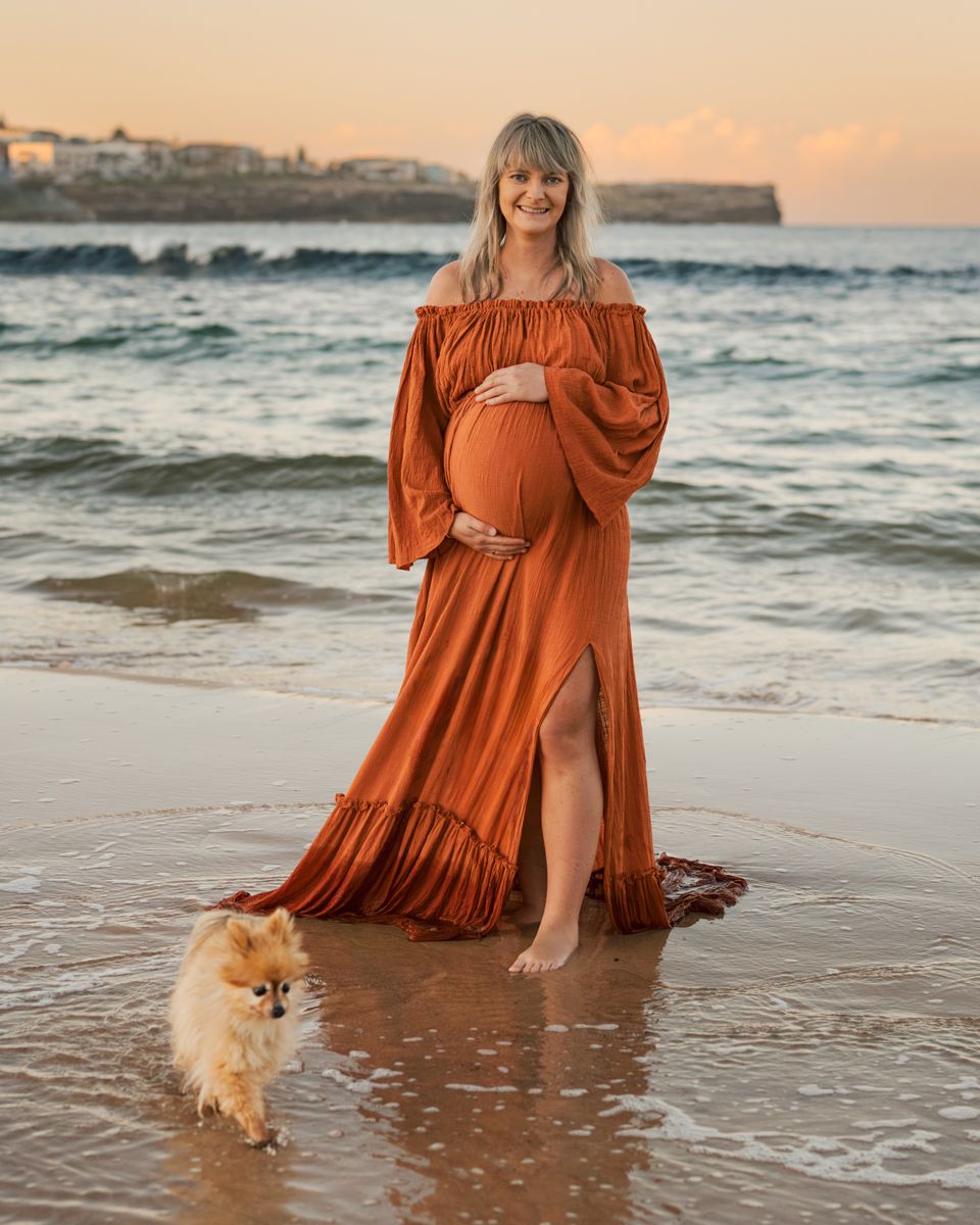 A person in a flowing rust-colored dress stands at the beach with a small Pomeranian dog during sunset.