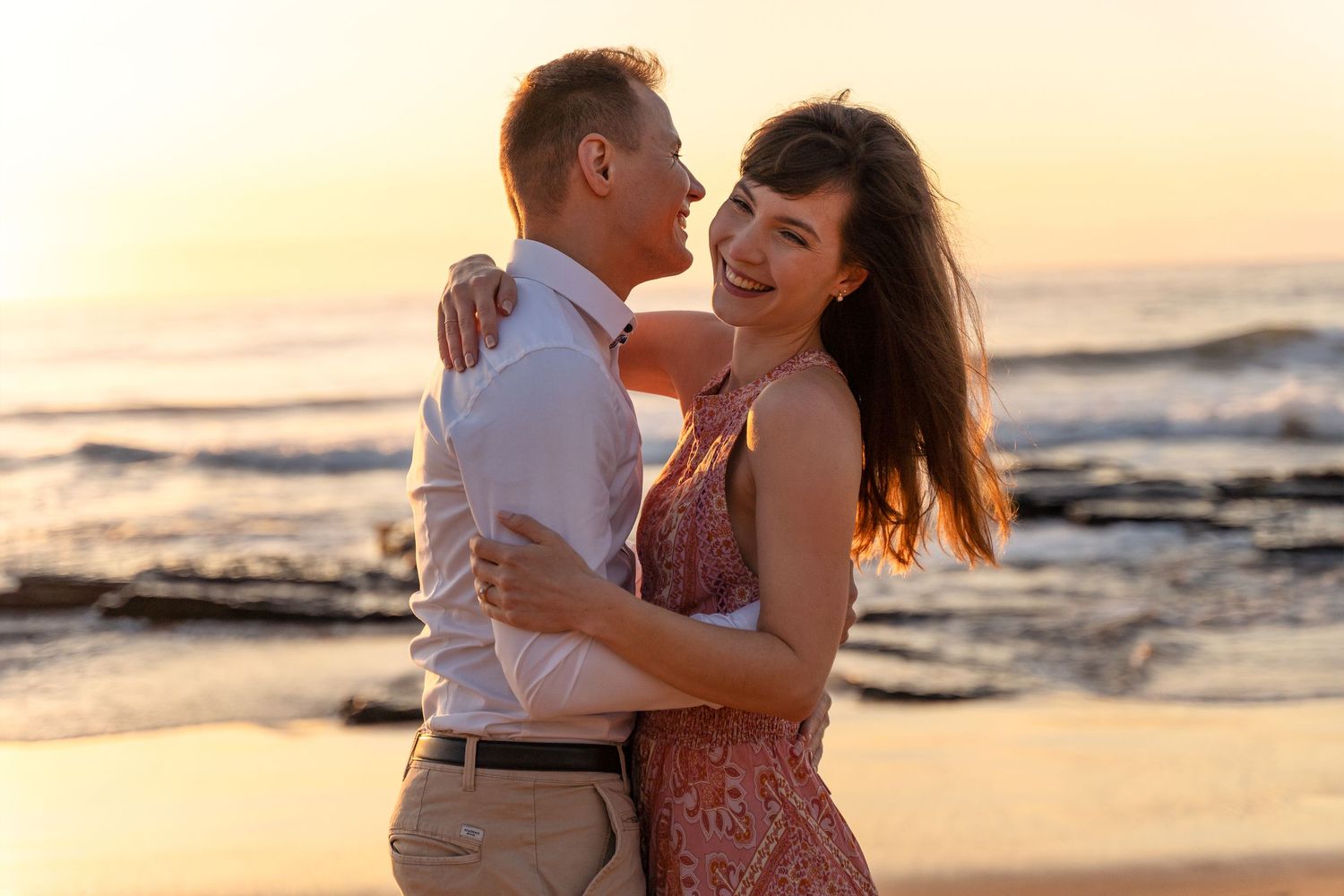 A couple hugging and laughing together on a beach at sunrise with ocean waves in the background captured by a couples photographer in Sydney