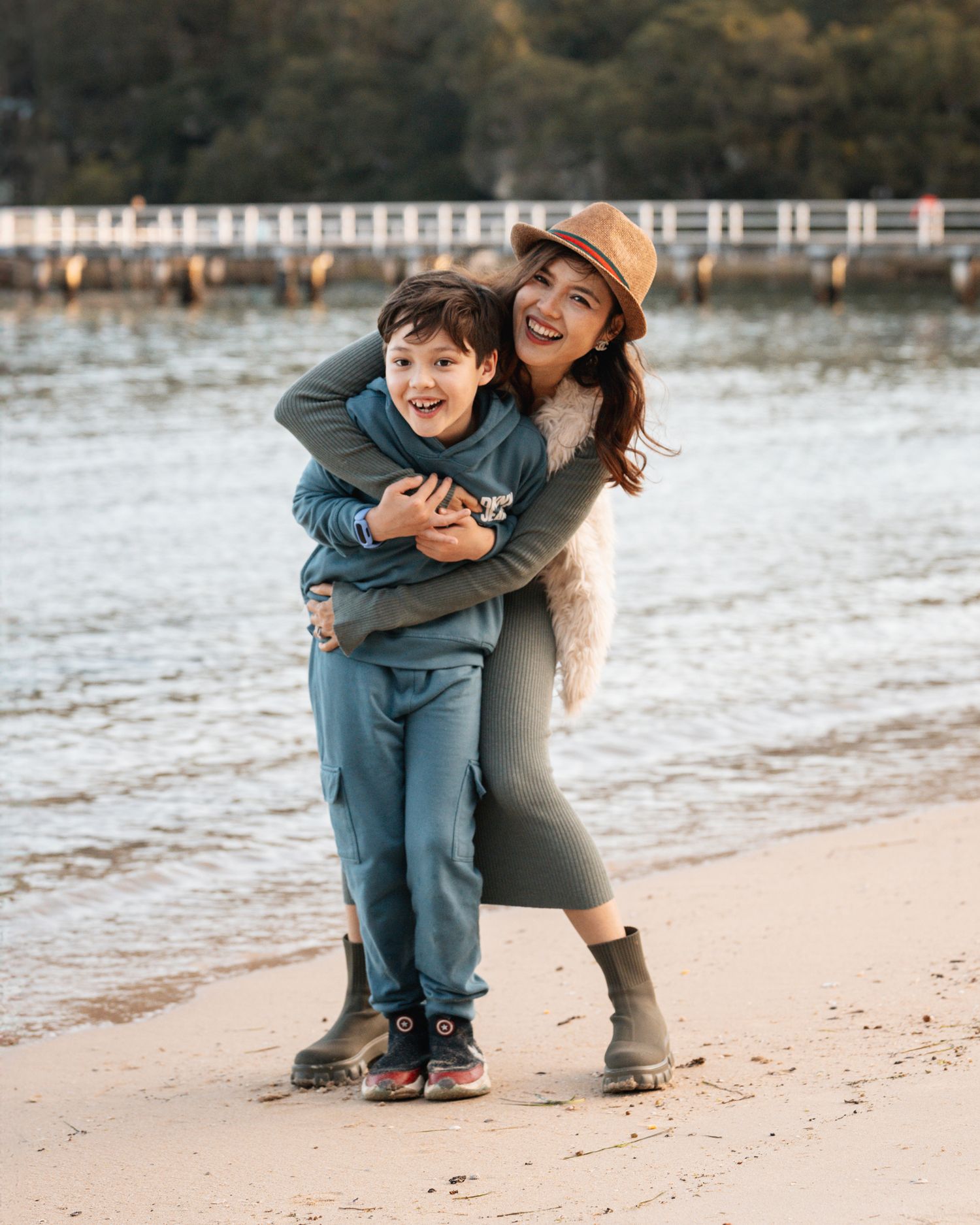 Mom and son share a joyful moment together on a sandy beach near a white fence on an overcast day in Clifton garden Sydney photoshoot