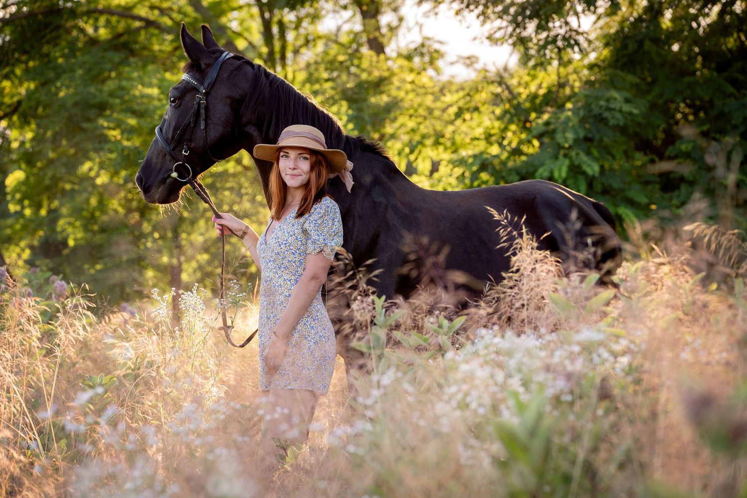 A black horse and girl in a flowing dress stand among tall wildflowers during golden hour in a meadow. Photoshoot with a horse in Cattai