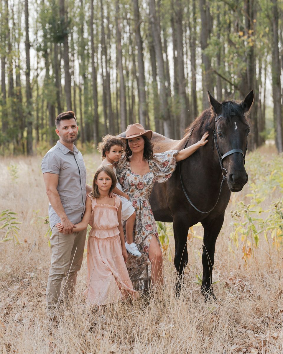 A family stands together with their black horse in a sunlit field of tall grass.