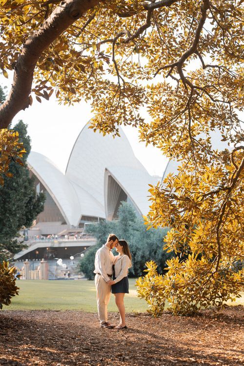 A couple embraces under golden autumn leaves with the iconic Sydney Opera House visible in the background. Proposal photoshoot in Sydney