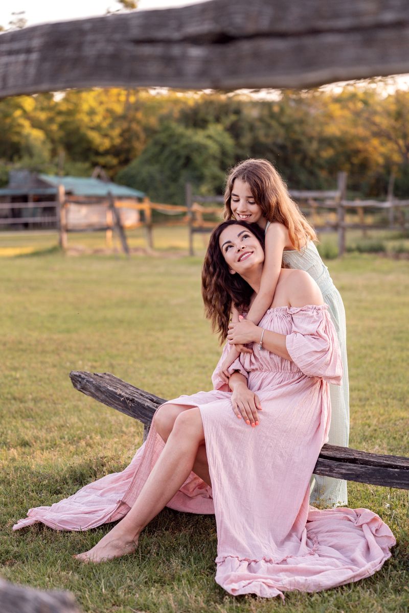 mom and daughter in a flowing pink dress sits gracefully on a rustic wooden fence in a sunlit meadow during golden hour photoshoot in sydney