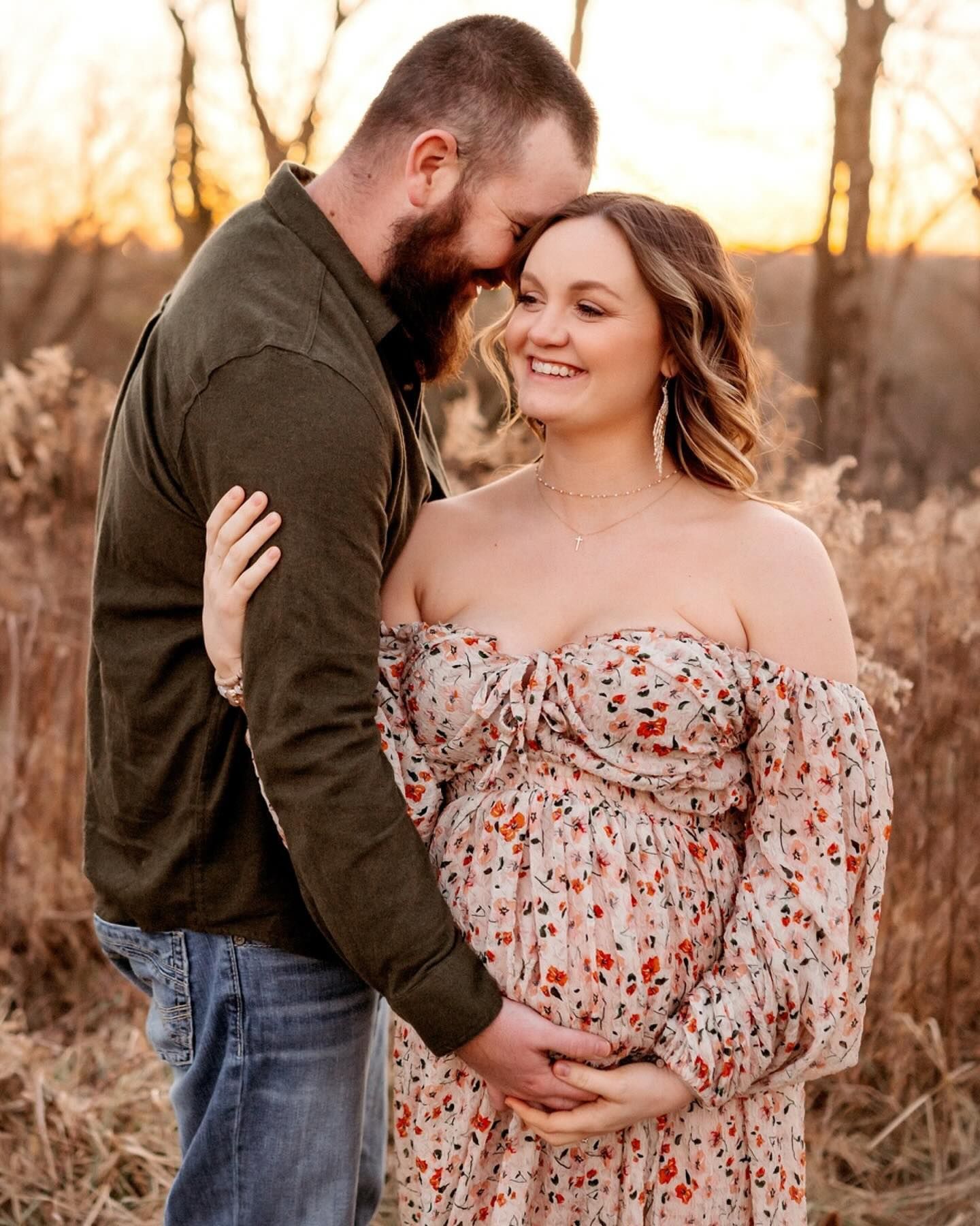 A couple shares an intimate moment at sunset in a field of tall grass wearing a floral dress and dark jacket.