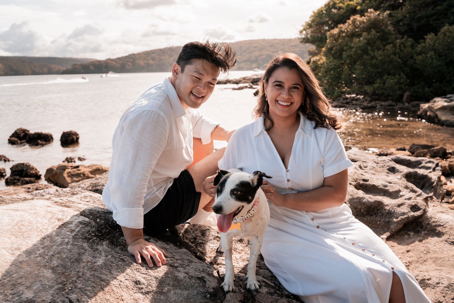 A couple in white outfits poses with their dog on a rocky beach during a sunset photoshoot in the northern beaches