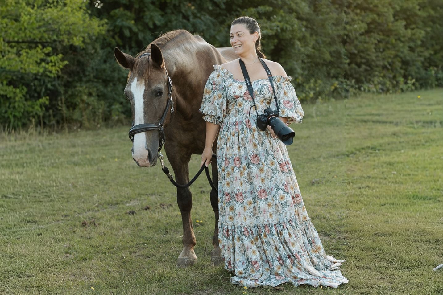 A person in a floral dress walks through a grassy field with a brown horse following behind them.