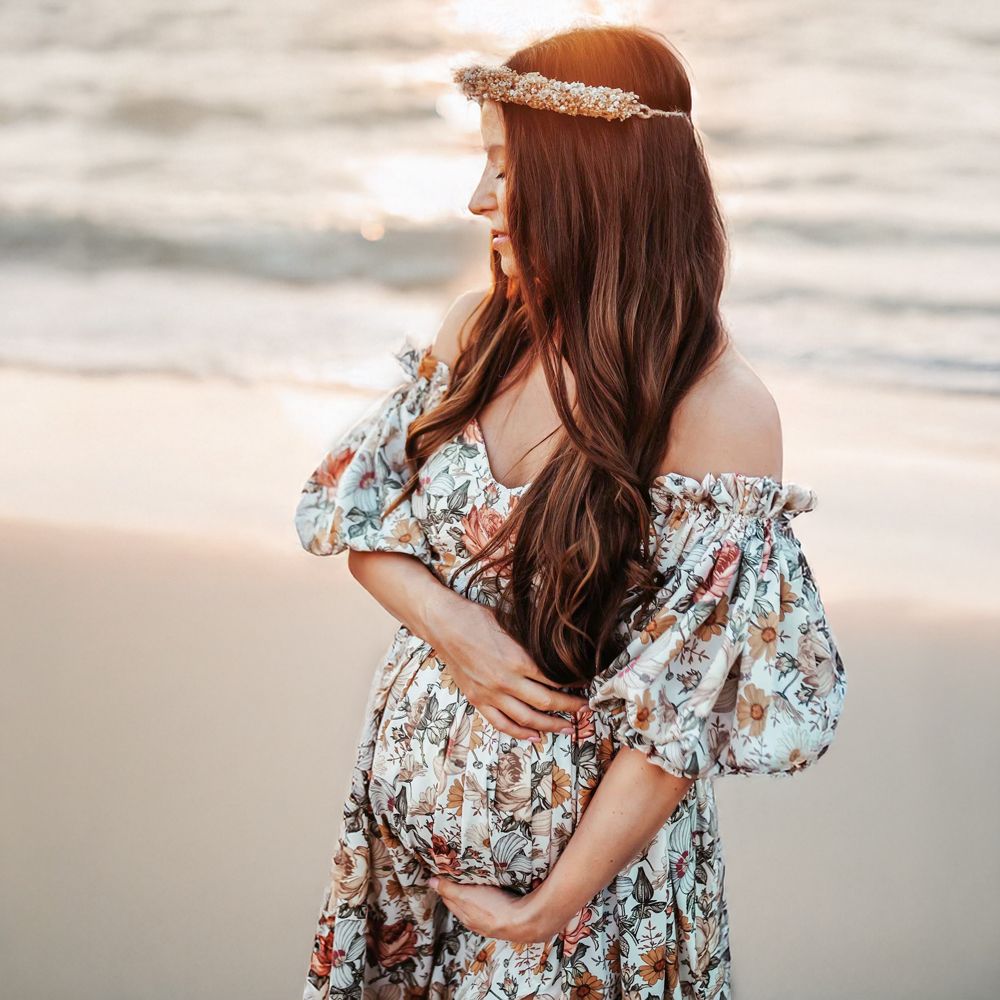 Woman in floral dress with ruffled sleeves embraces herself on a misty beach at sunset.