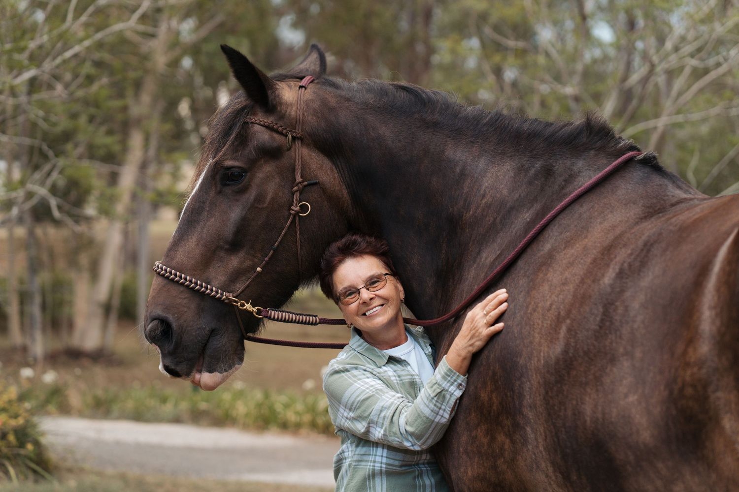 Woman in plaid shirt smiling beside a dark brown horse with bridle outdoors.