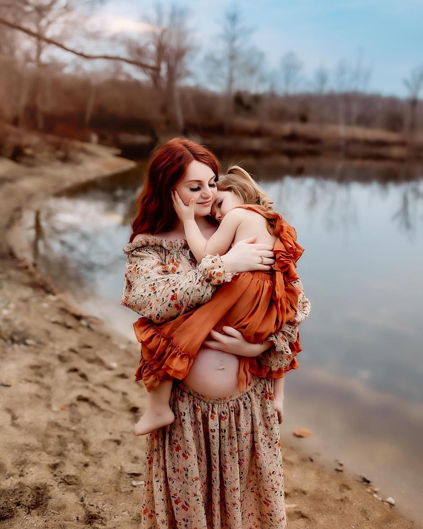 A tender moment captured by a lake with earth-toned clothing and autumn scenery in the background.