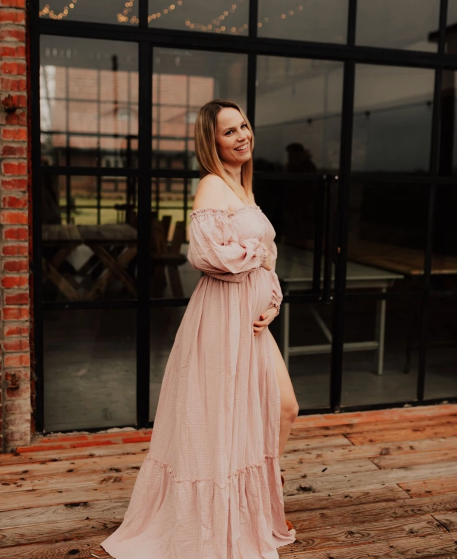 A woman in a flowing pink maxi dress poses gracefully against an industrial brick wall with string lights.