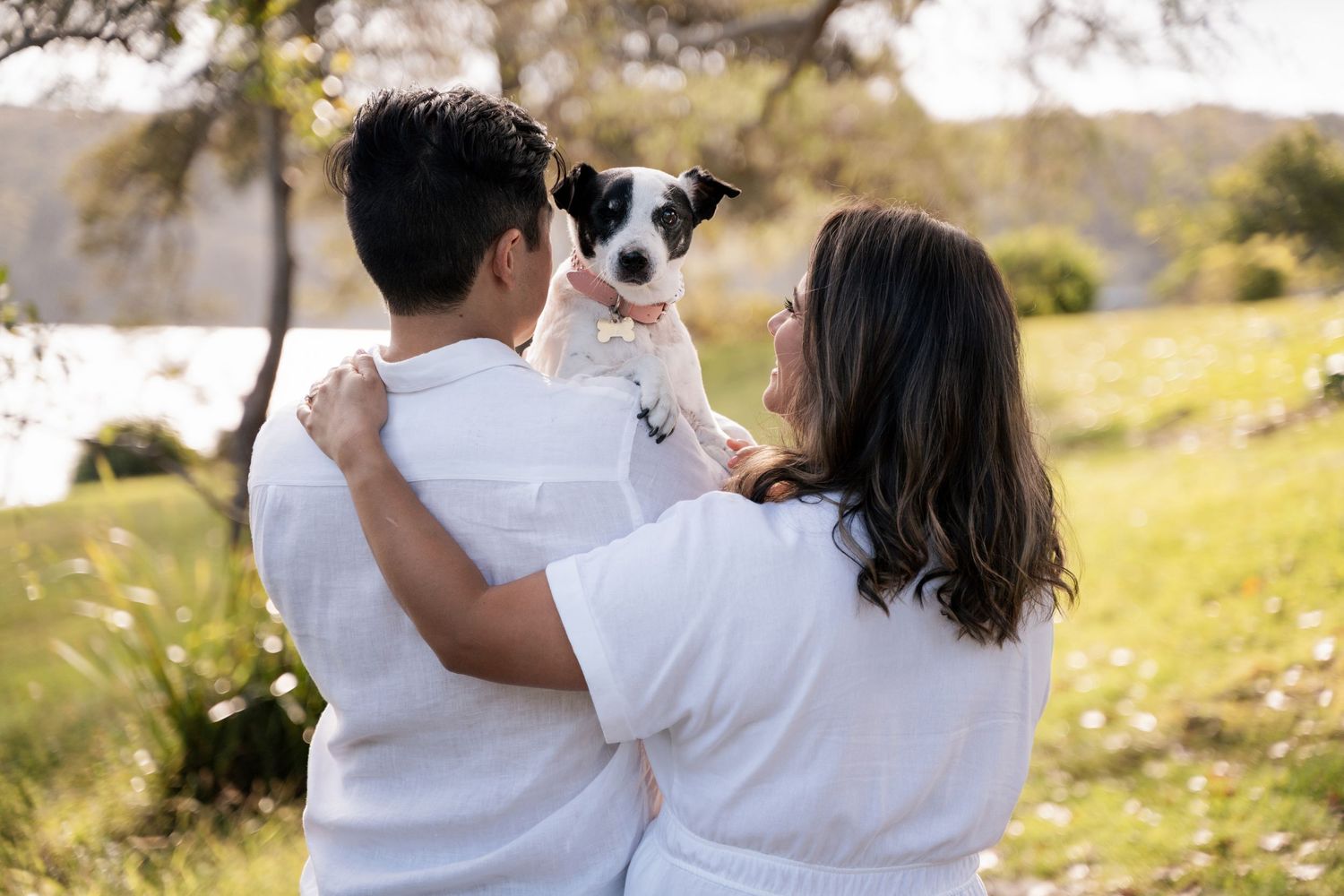 A couple in white shirts holds their small black and white dog while sitting in a sunny meadow during a photoshoot with dog