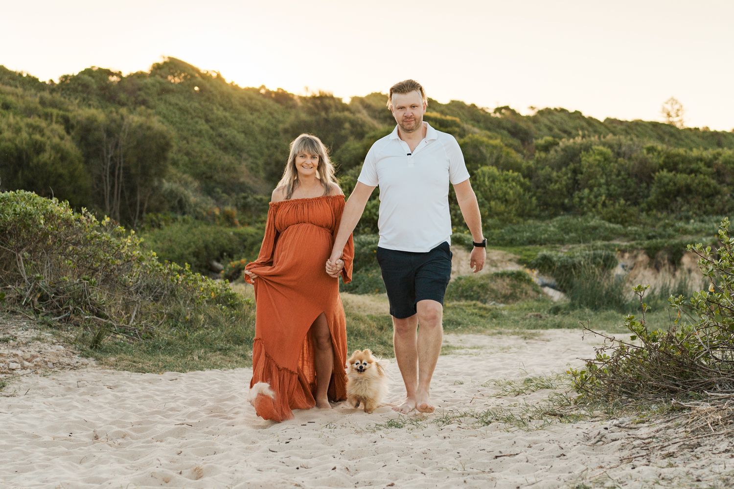A couple walks along a sandy beach path in golden hour with natural surrounding them during a maternity photoshoot in Rouse Hill