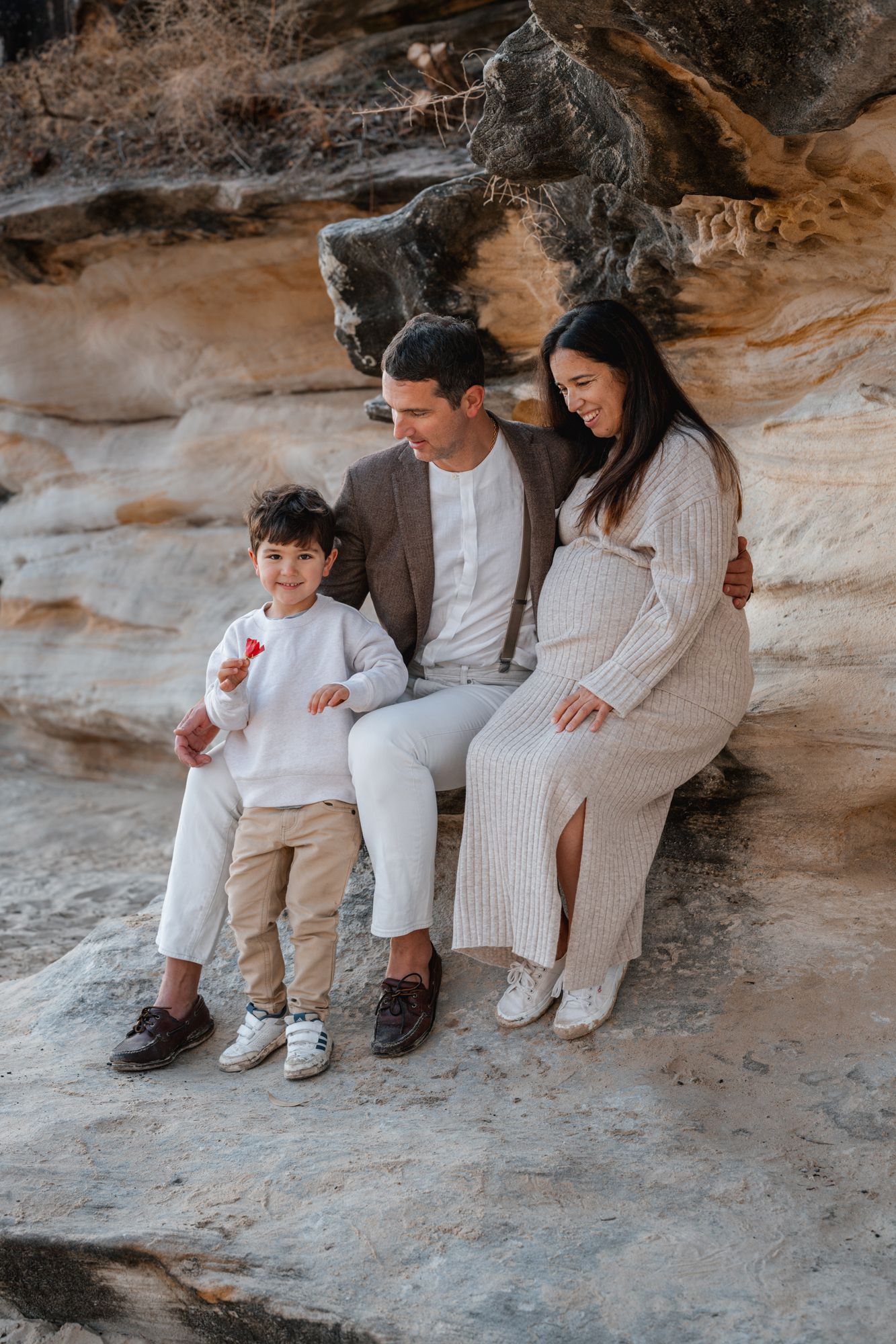 A family in neutral-colored clothing sits together on a rocky ledge in a natural outdoor setting.