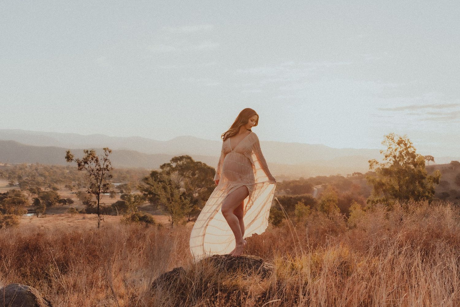 Woman in flowing dress standing on hilltop against desert mountain landscape at golden hour.