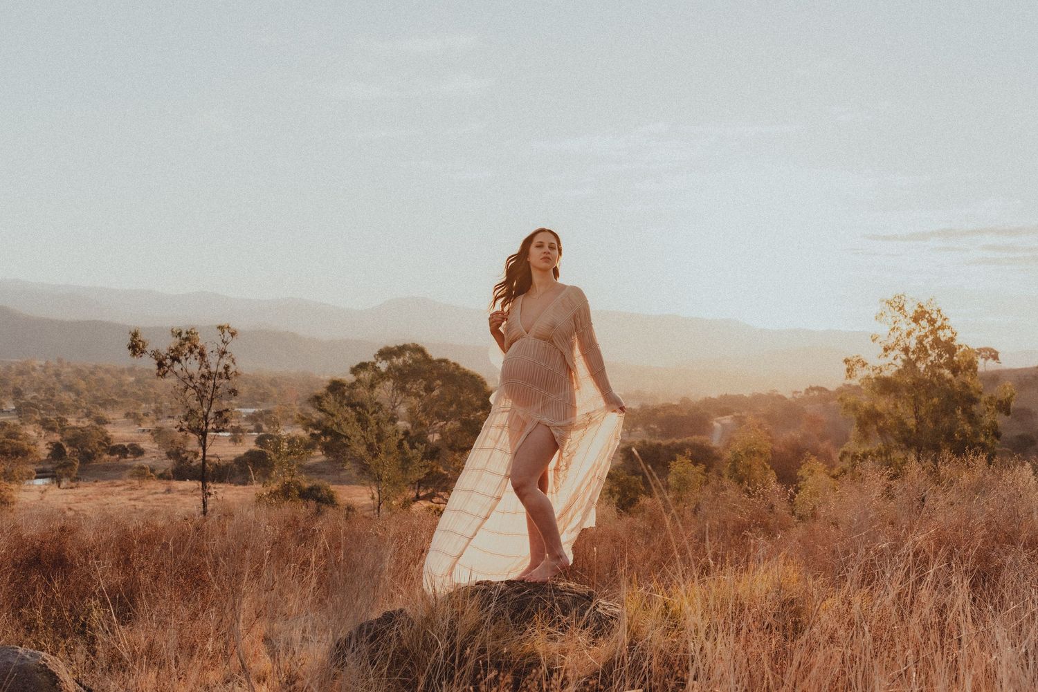 Woman in flowing dress walking through golden grass with mountains in background.