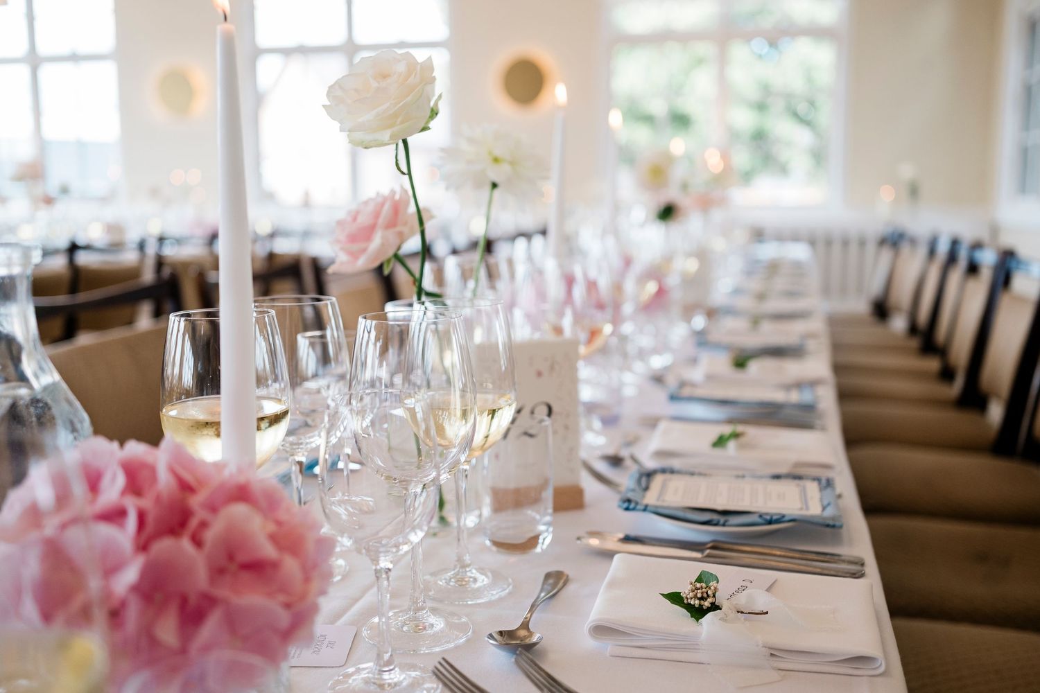 Elegant wedding reception table setting with pink hydrangeas, white roses, crystal glasses, and candles on white linen.