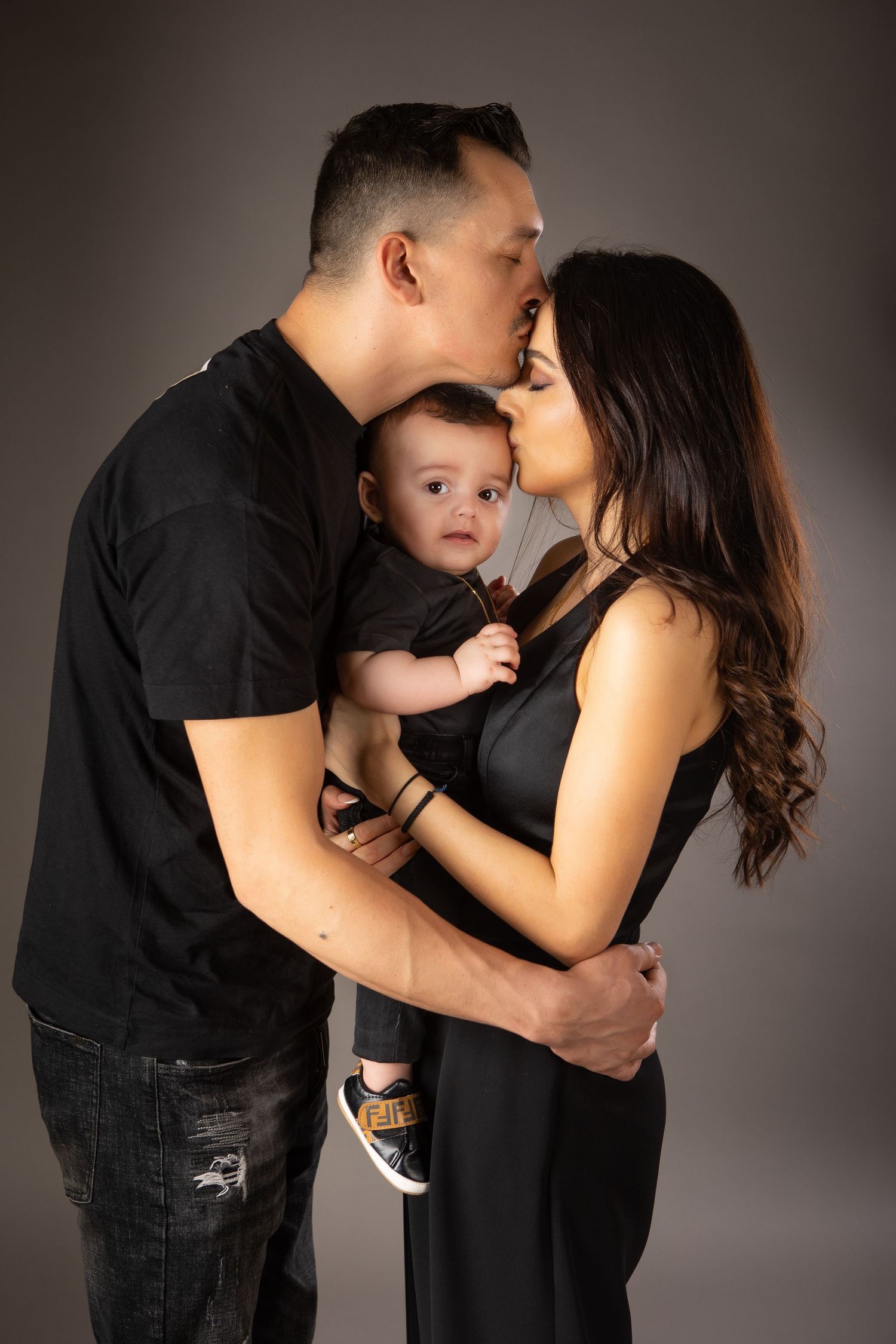 mum and dad are holding their little son together while the dad is kissing the mum on her forehead. the baby is looking at the camera
