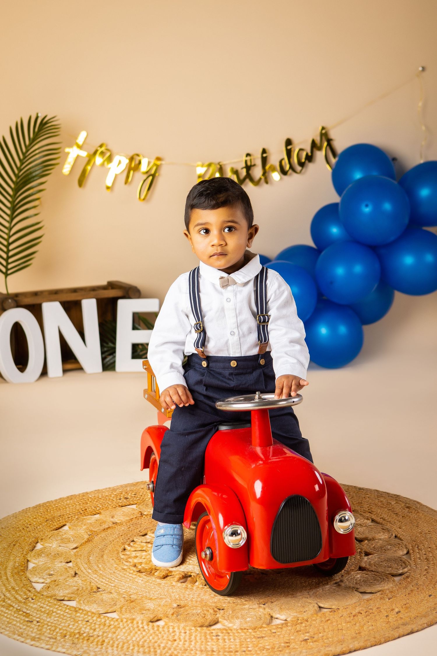 A one-year-old boy is sitting on a red fire engine metal car with  blue balloons and the banner Happy Birthday in the background