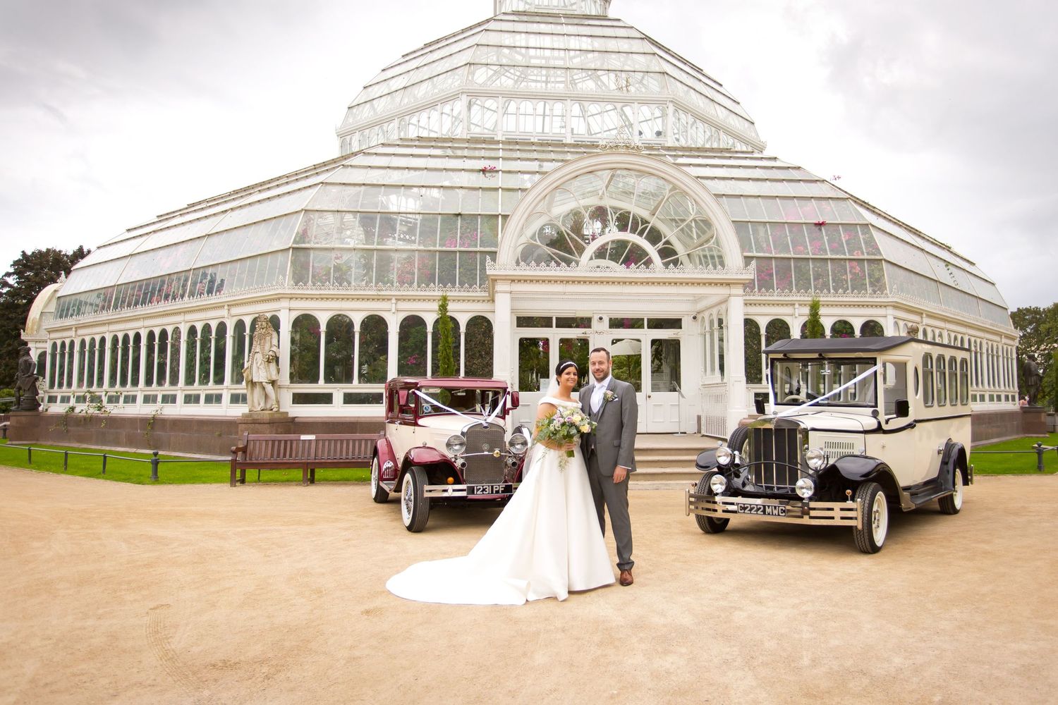 The bride and groom are posing in front of the wedding cars in Sefton Park with Palm House at the back