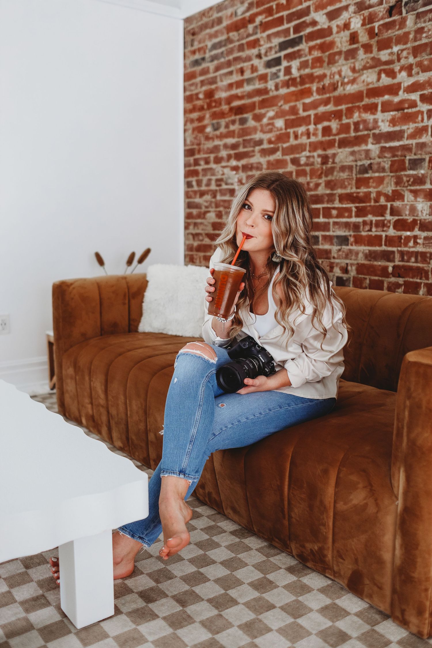 Woman in casual attire relaxes on brown velvet couch against brick wall while holding a cold beverage.