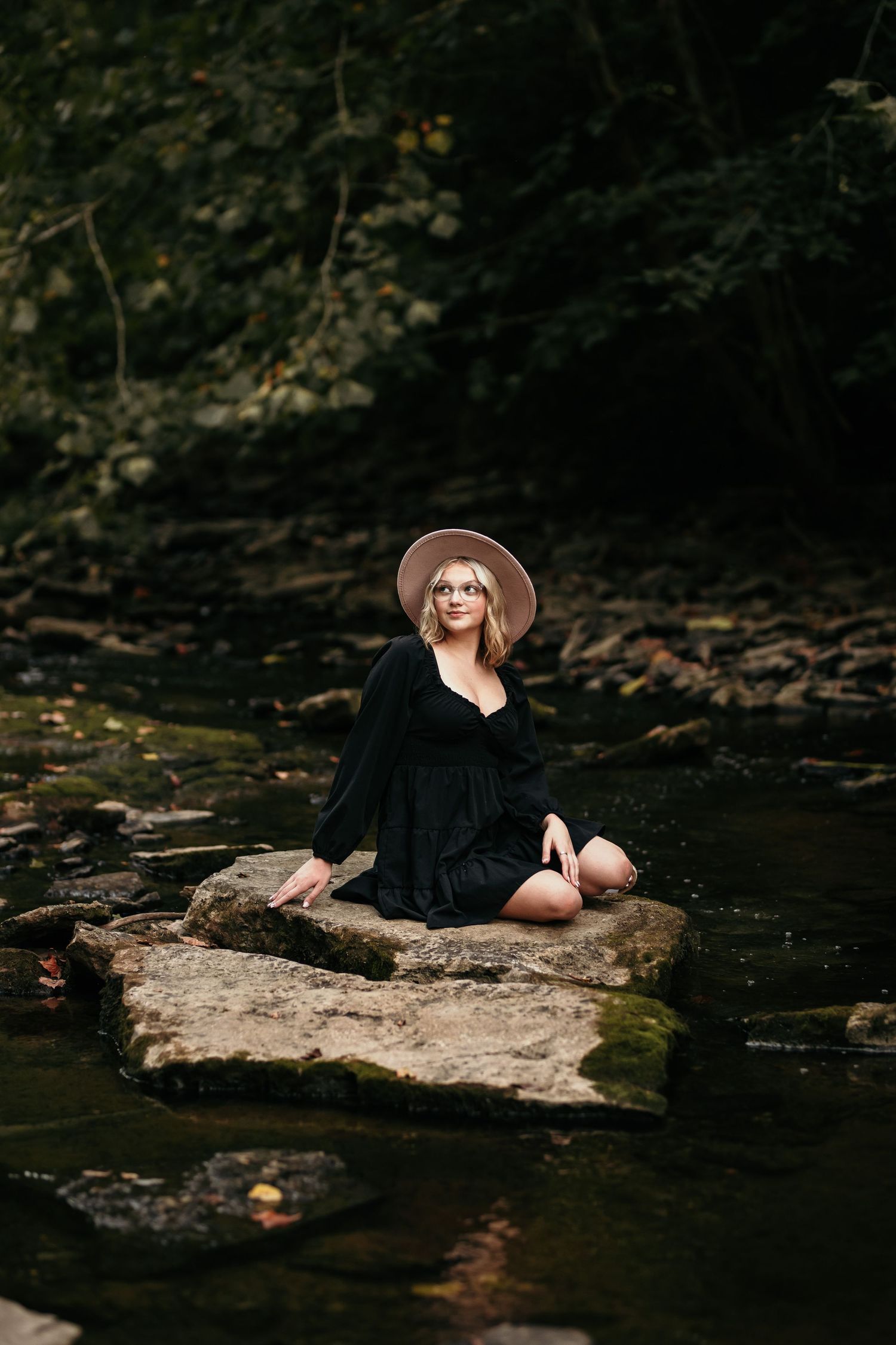 Woman in black dress and hat sitting on rock by stream in moody forest setting.