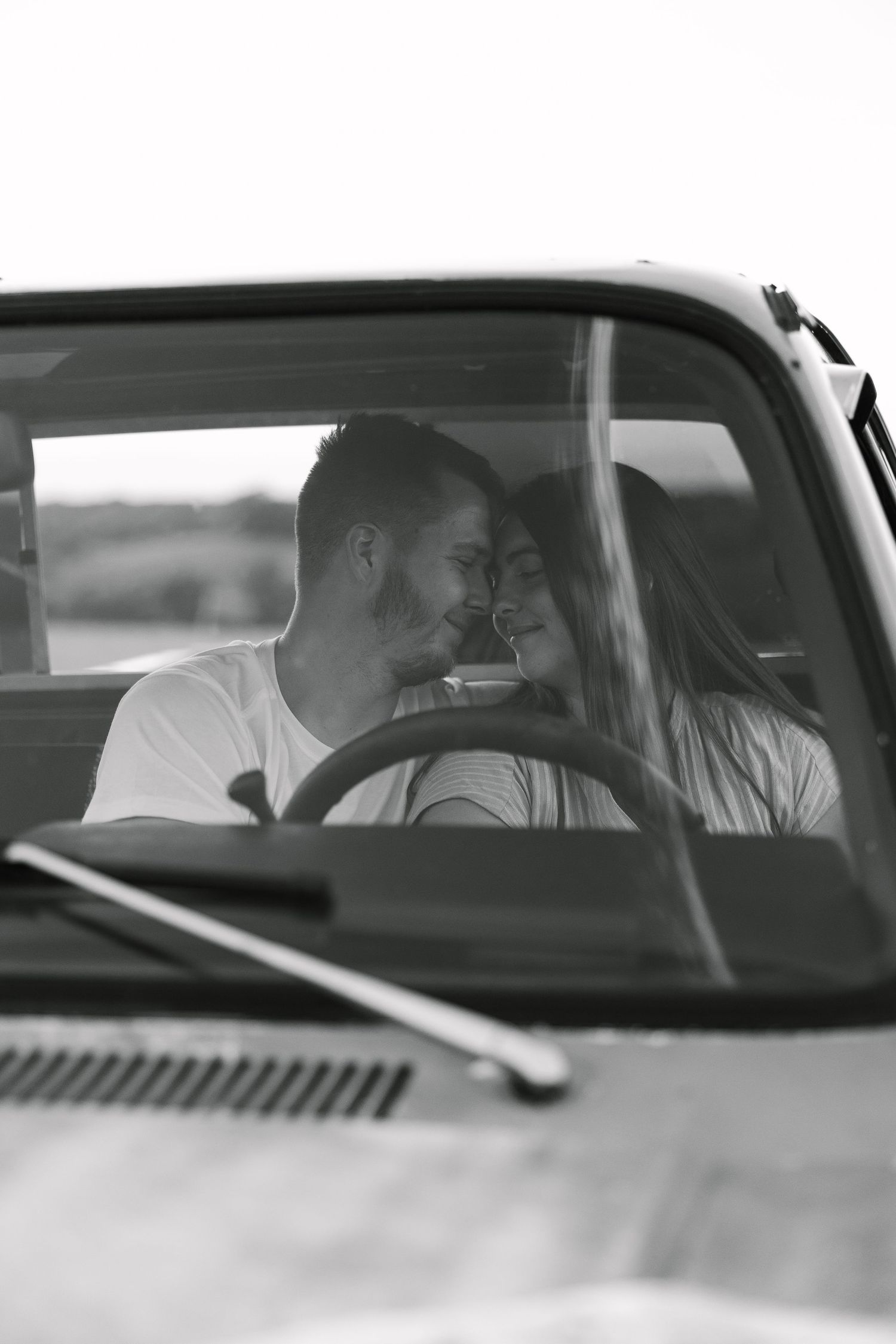 Black and white photo of a romantic couple sitting close inside a vintage truck, touching foreheads and smiling through the windshield.