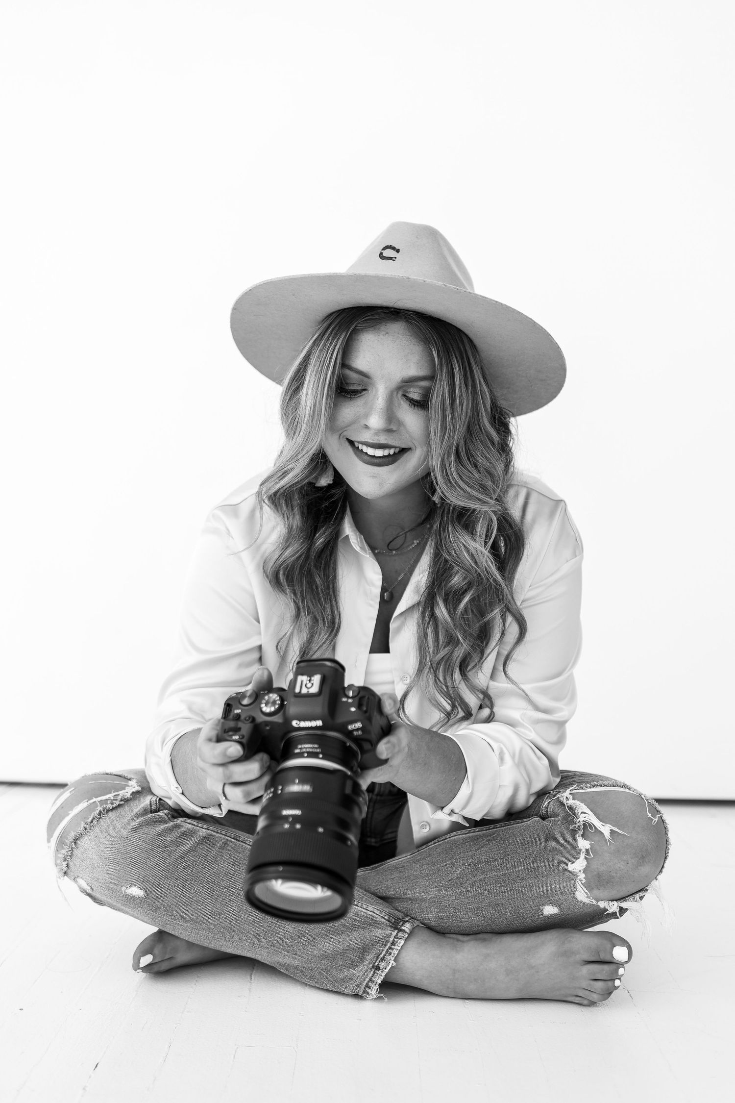 Photographer sitting cross-legged on white floor wearing fedora hat and holding professional camera.