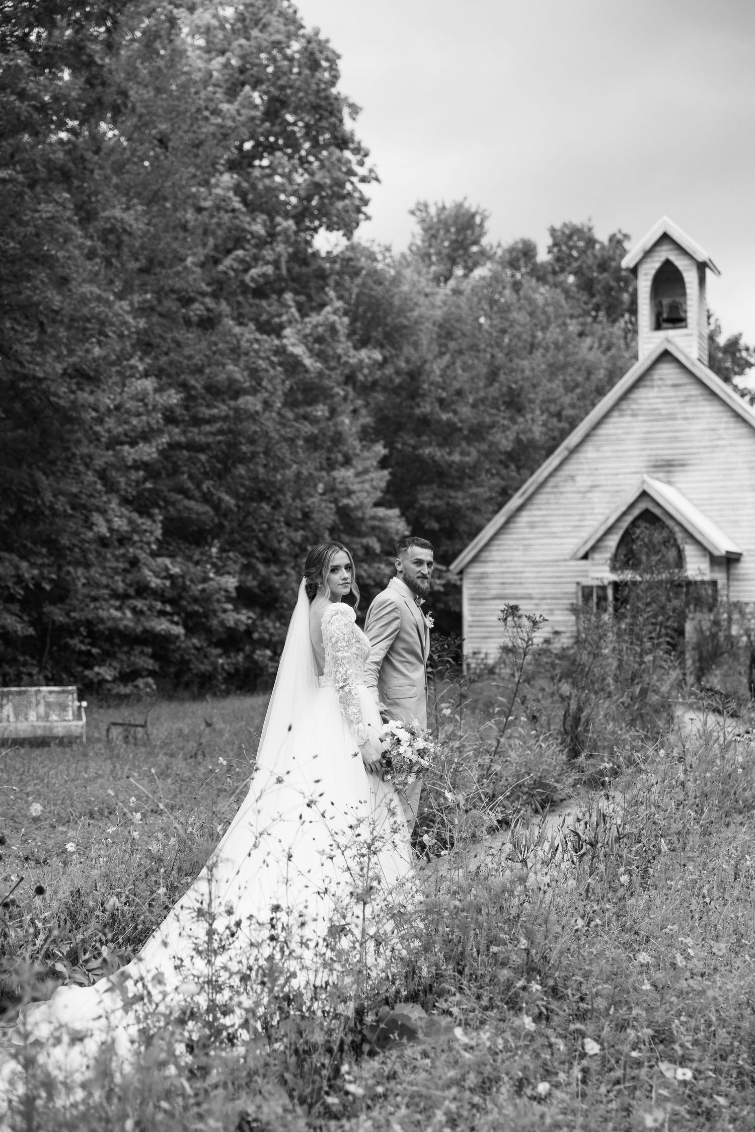 A black and white wedding portrait beside a charming country chapel surrounded by wildflowers.