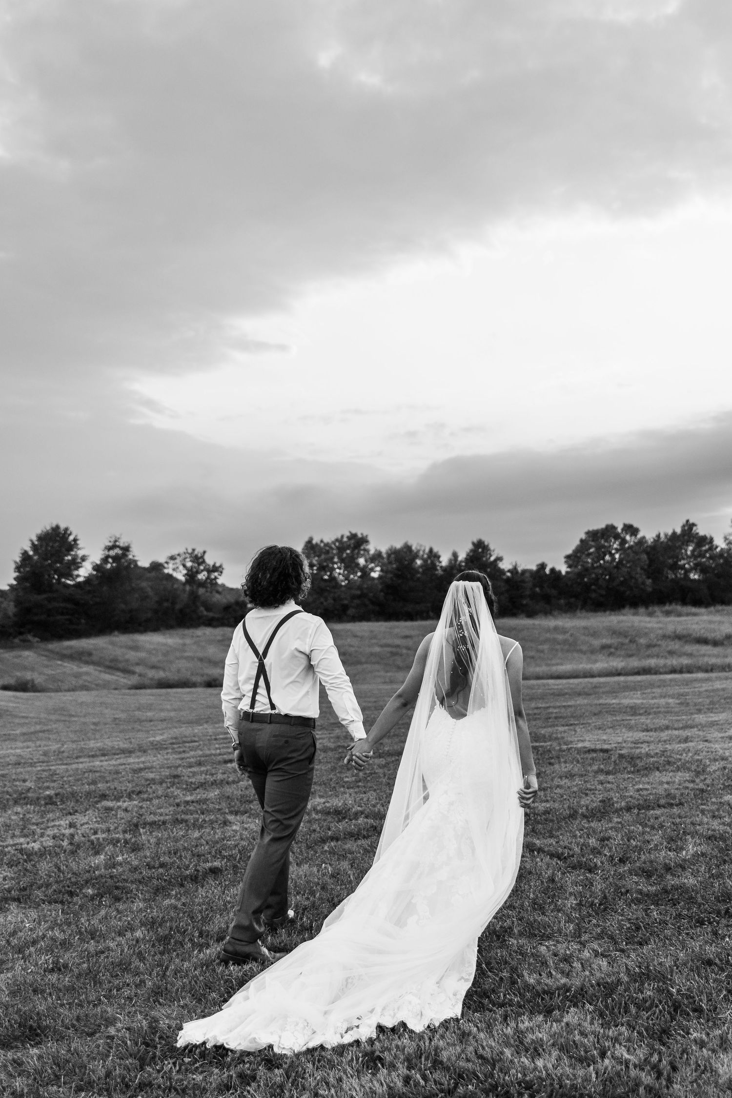 A couple walking away across a meadow, the bride's white dress trailing behind them.