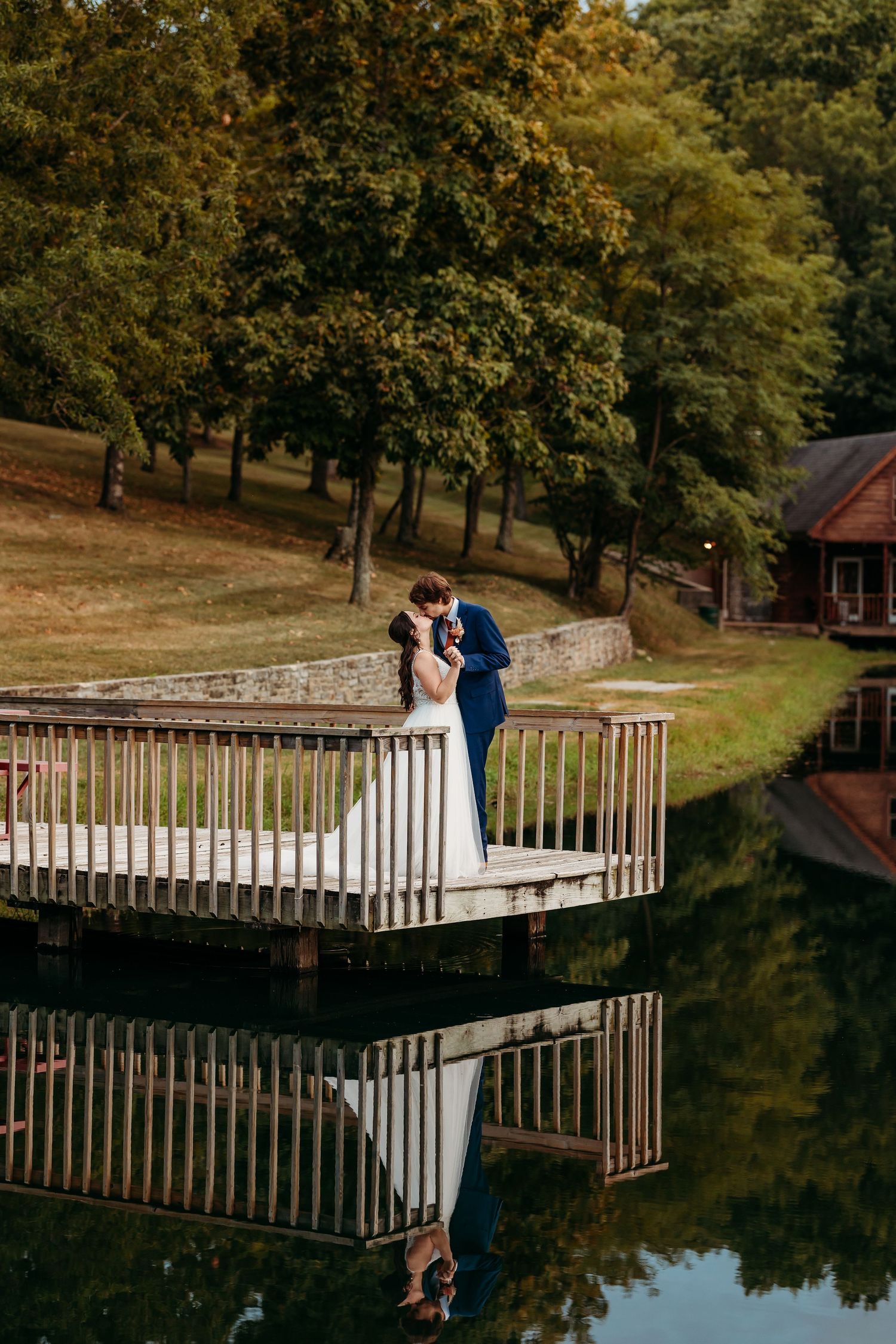 A couple shares a romantic moment on a wooden dock overlooking a pond surrounded by autumn trees.