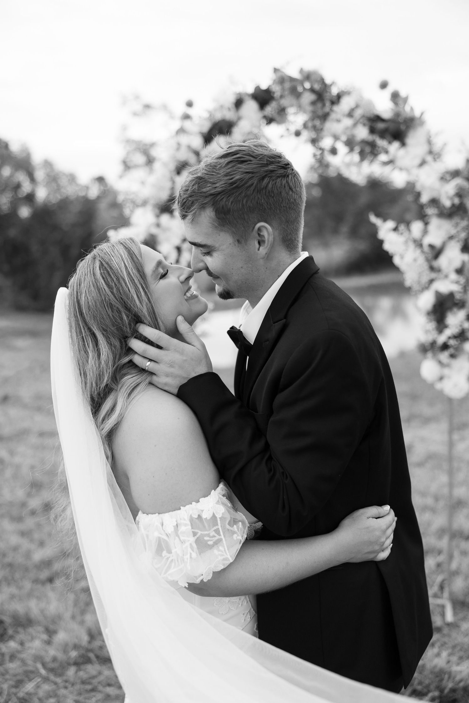 Black and white photo of a bride and groom sharing an intimate moment, nearly kissing, in front of a floral arch.