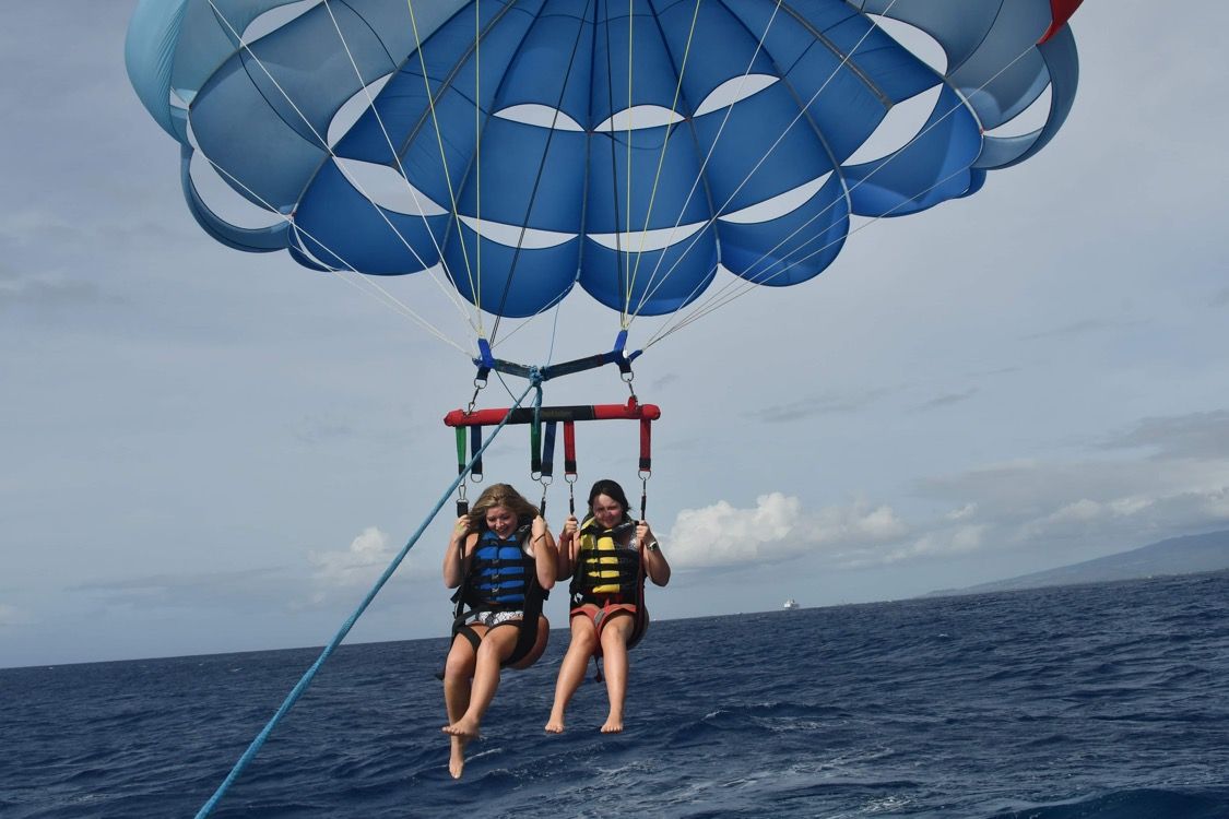 Two parasailers gliding over ocean waters with blue parachute against cloudy sky.