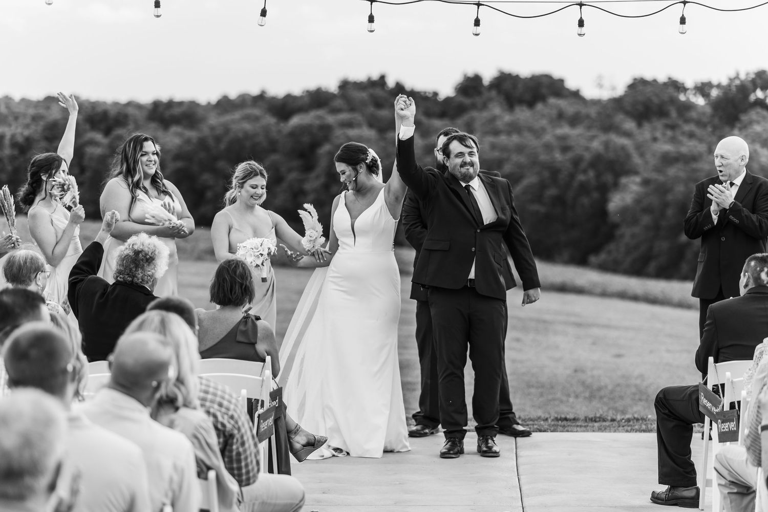 Newlyweds celebrate with raised arms while guests cheer during outdoor ceremony recessional.