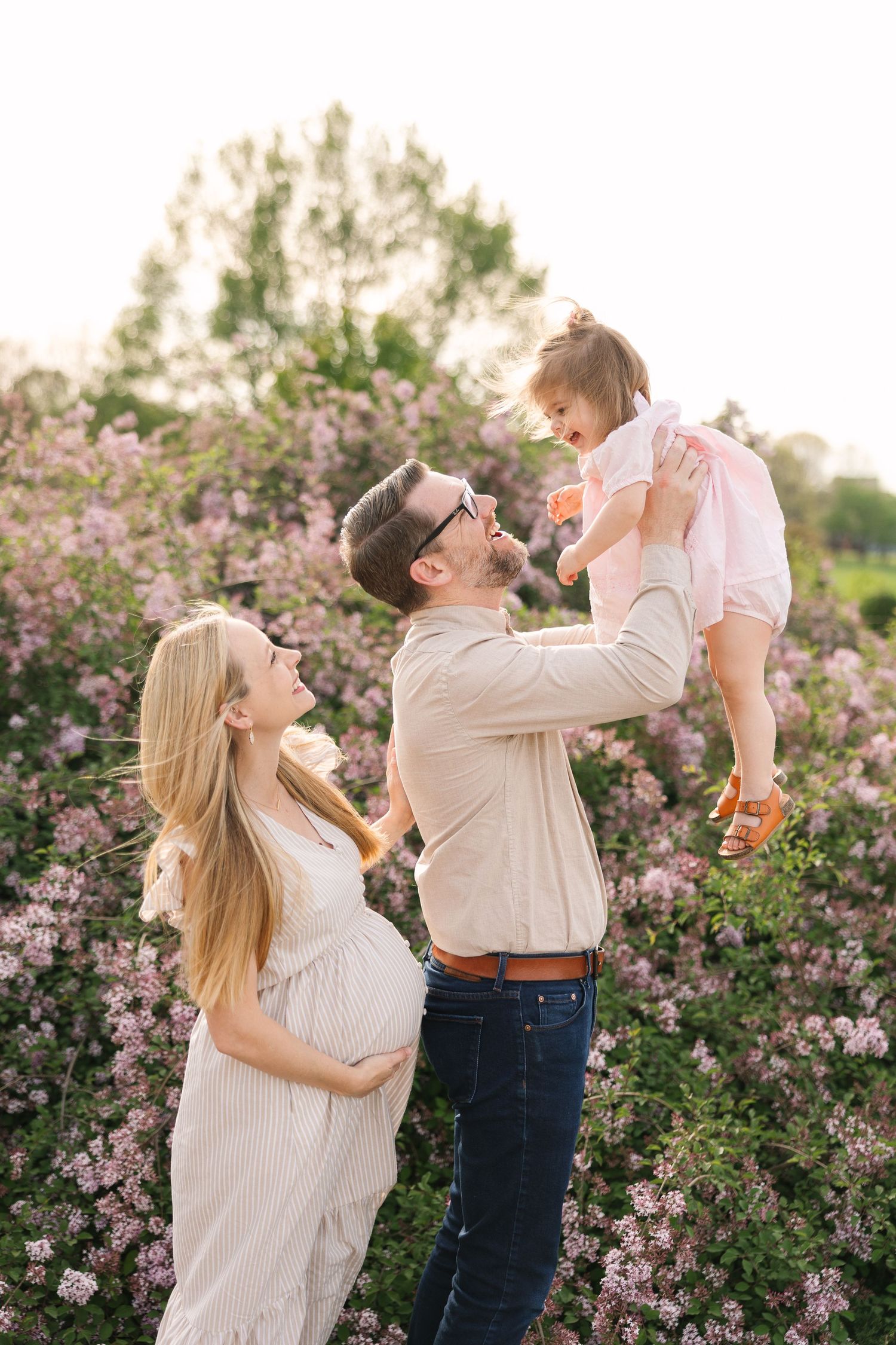 Pregnant woman in white dress with husband lifting toddler girl in pink dress among blooming lilac bushes outdoors.