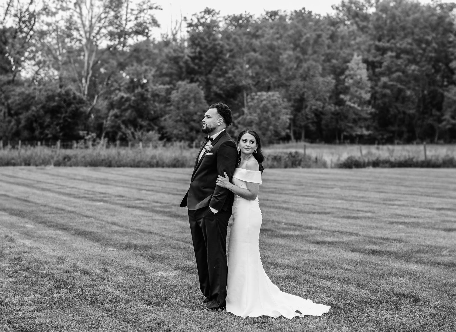 Black and white portrait of couple in formal attire standing back to back in field.