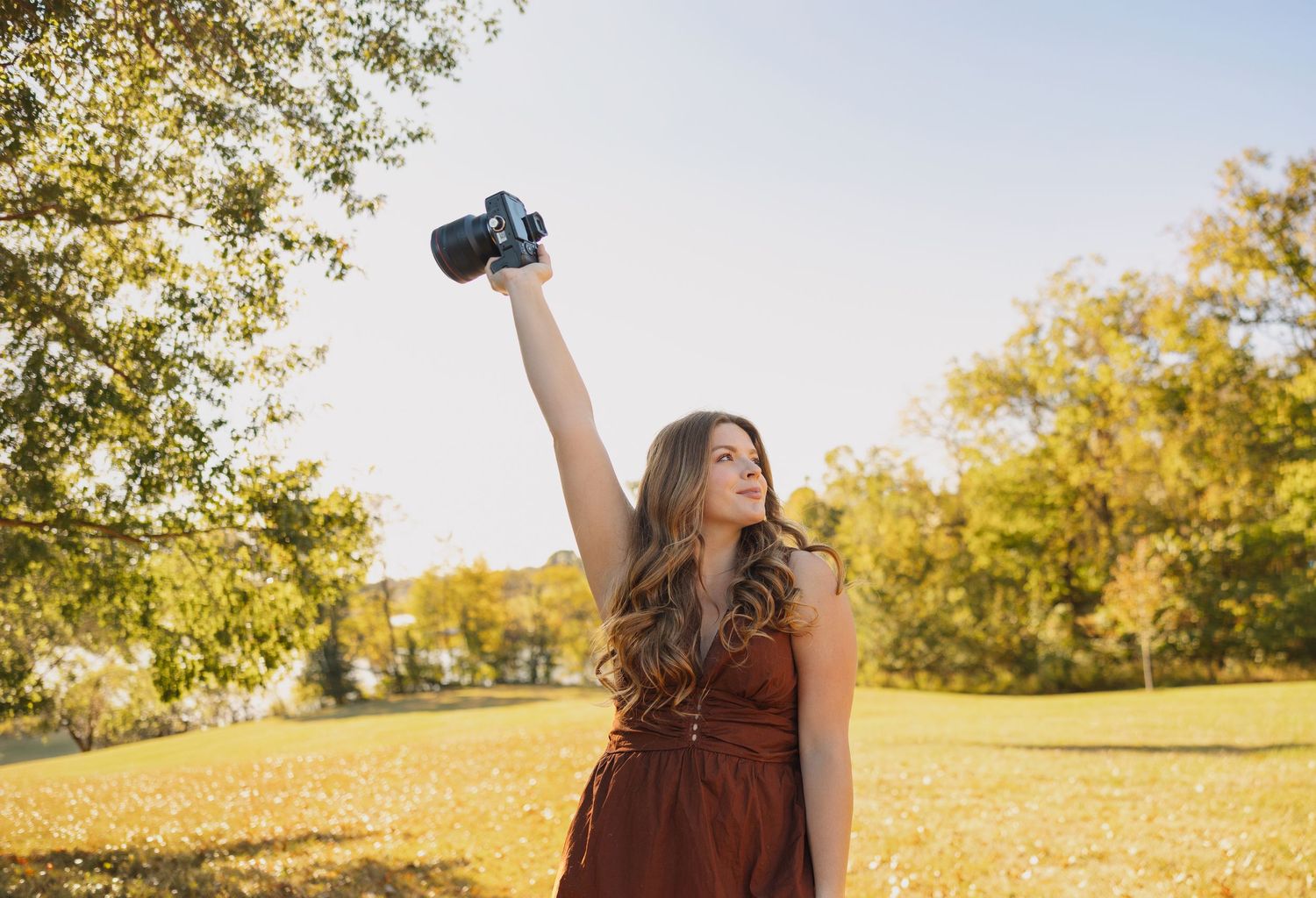 Young woman in red dress raises DSLR camera triumphantly in sunny autumn park with golden trees.