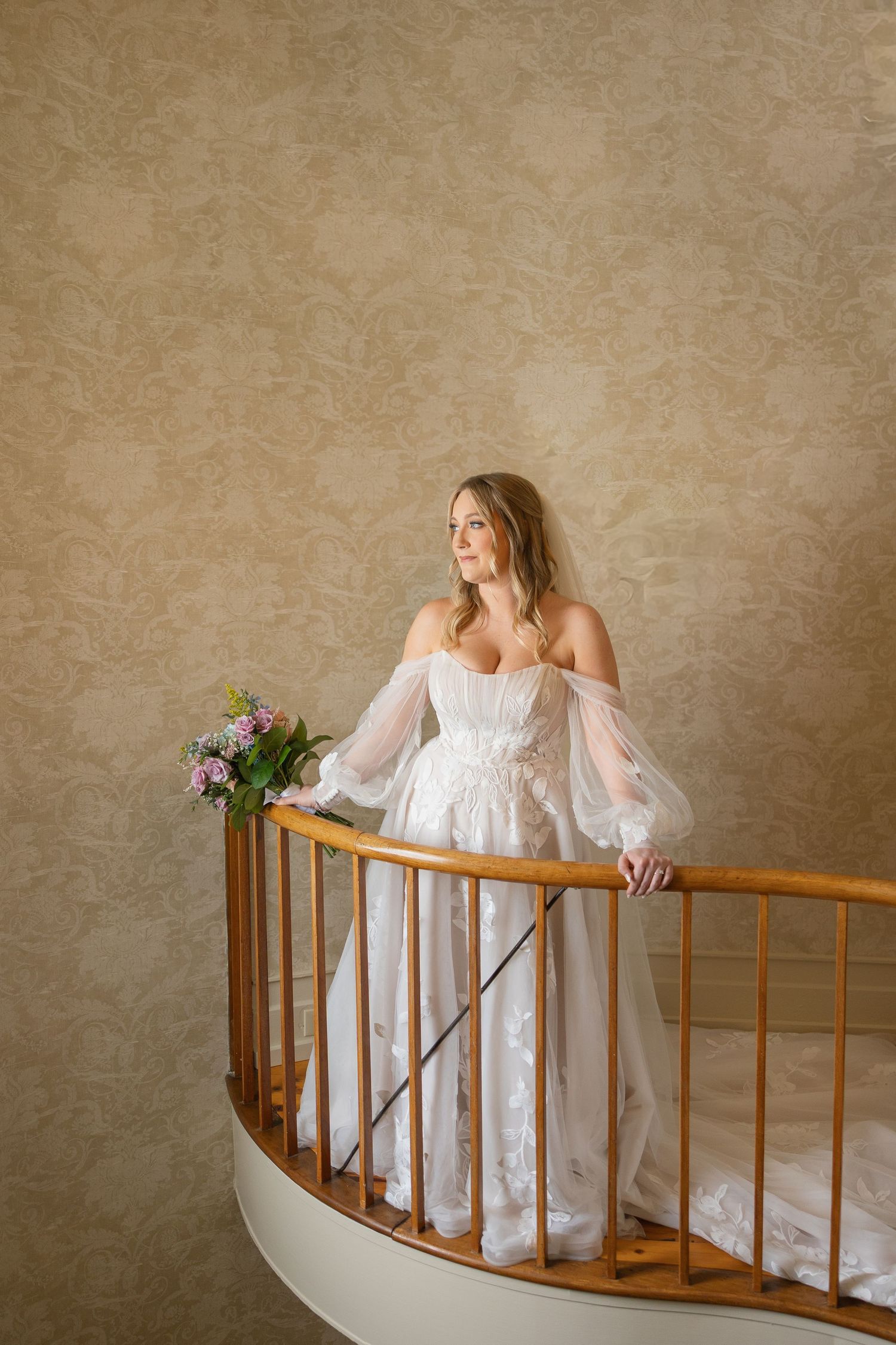 Bride in white off-shoulder gown with sheer sleeves holding floral bouquet on curved staircase.