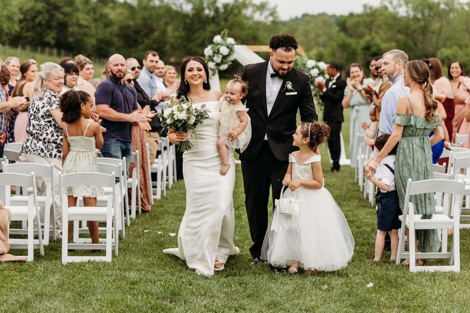 Wedding ceremony processional on grassy lawn with white chairs and guests looking on.