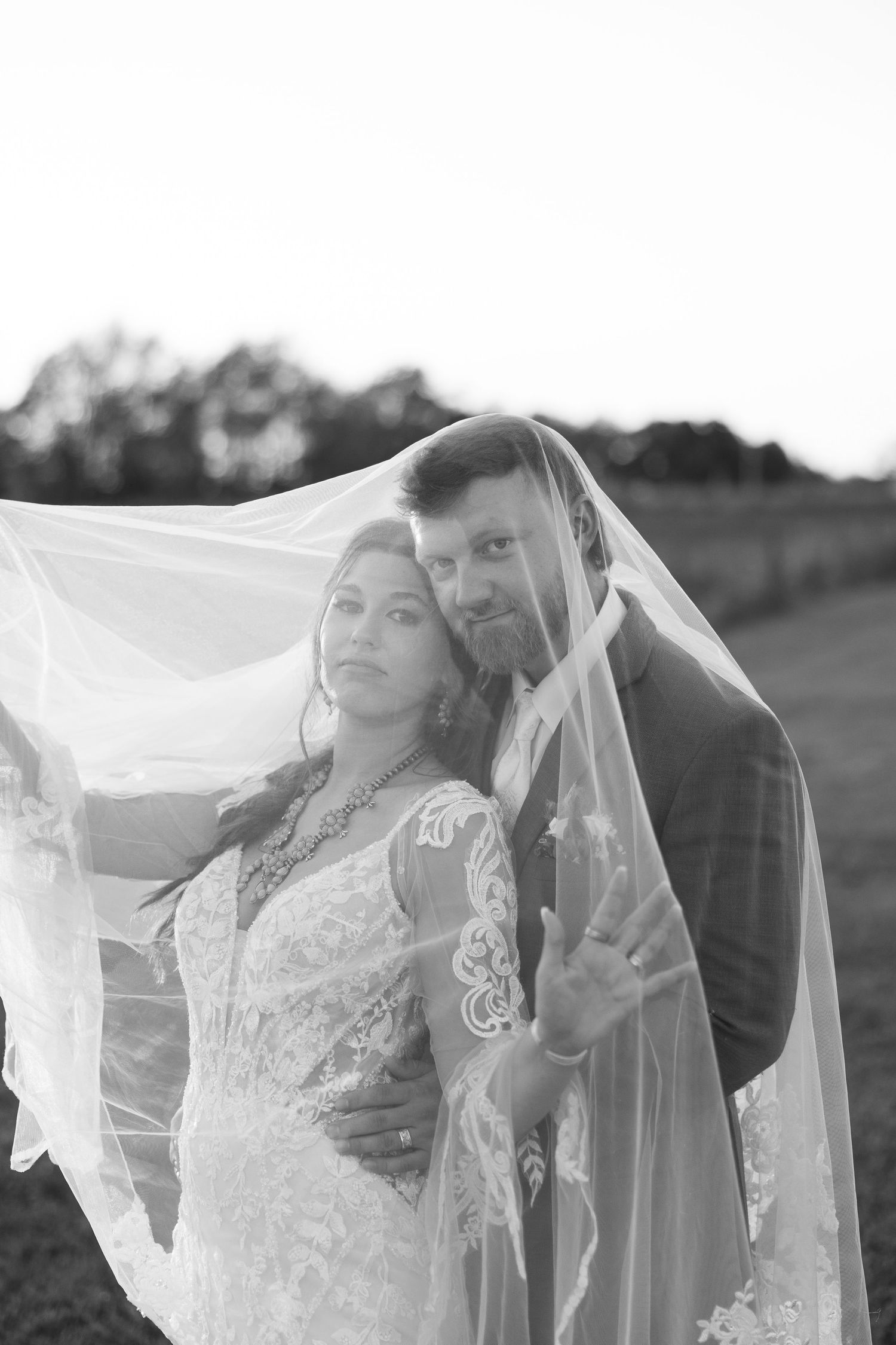 Black and white wedding portrait of a bride in a lace gown and a groom in a suit embracing beneath a sheer veil in an open field.