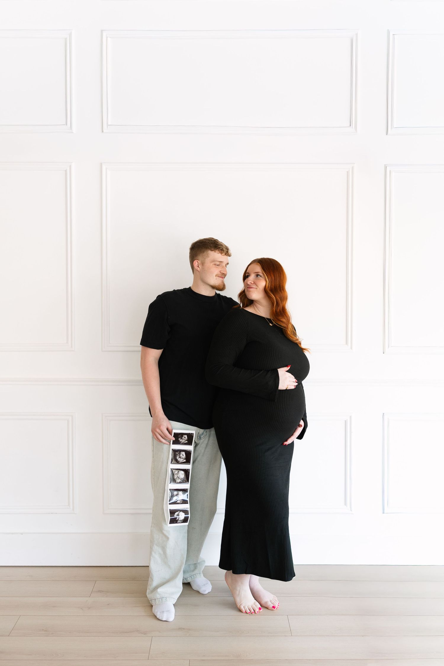 Expecting couple in black outfits pose in a white studio as the woman cradles her baby bump and the man holds an ultrasound photo strip.